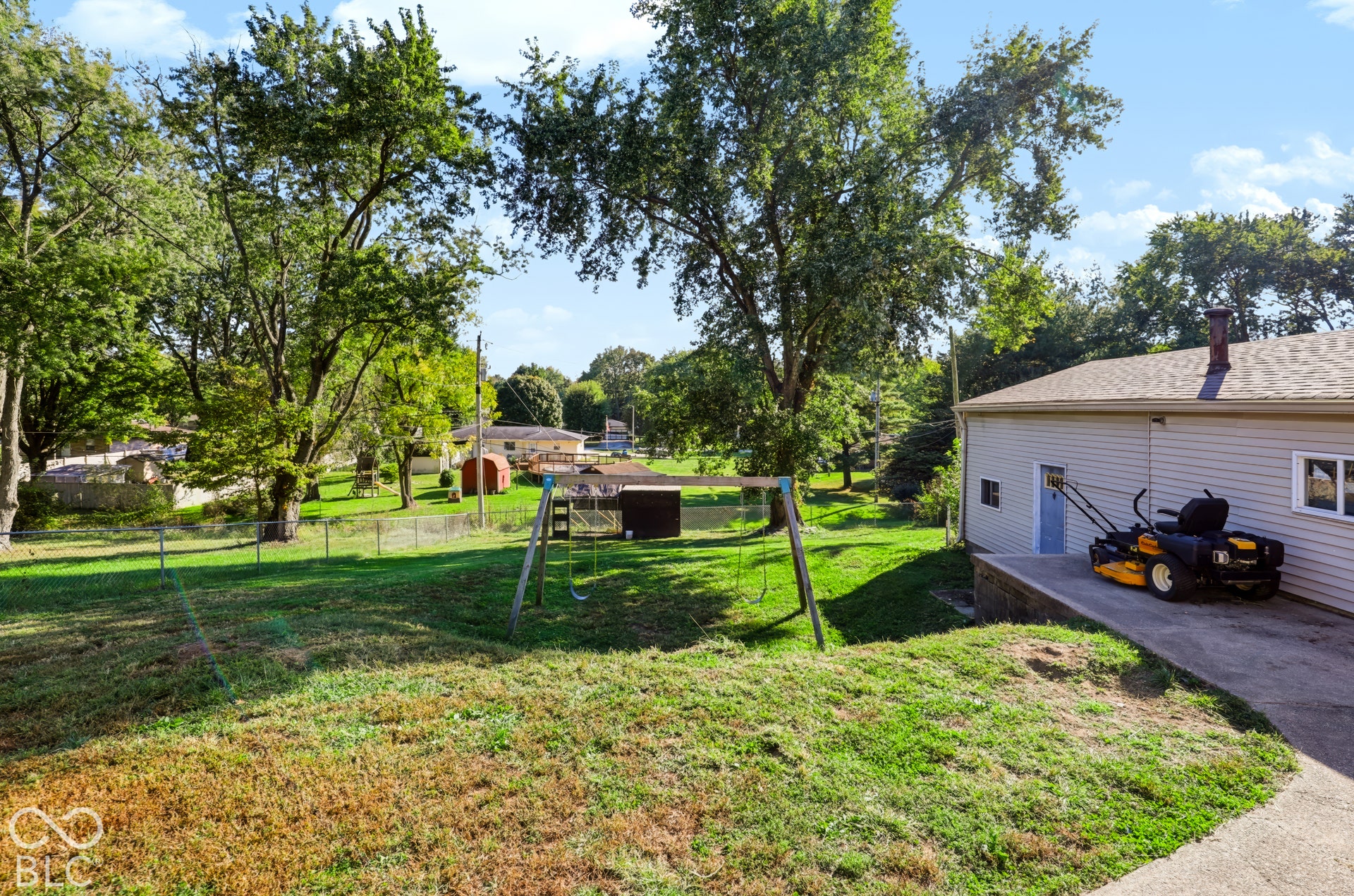 Expansive grass backyard featuring mature trees, a chain-link fence, and a concrete patio