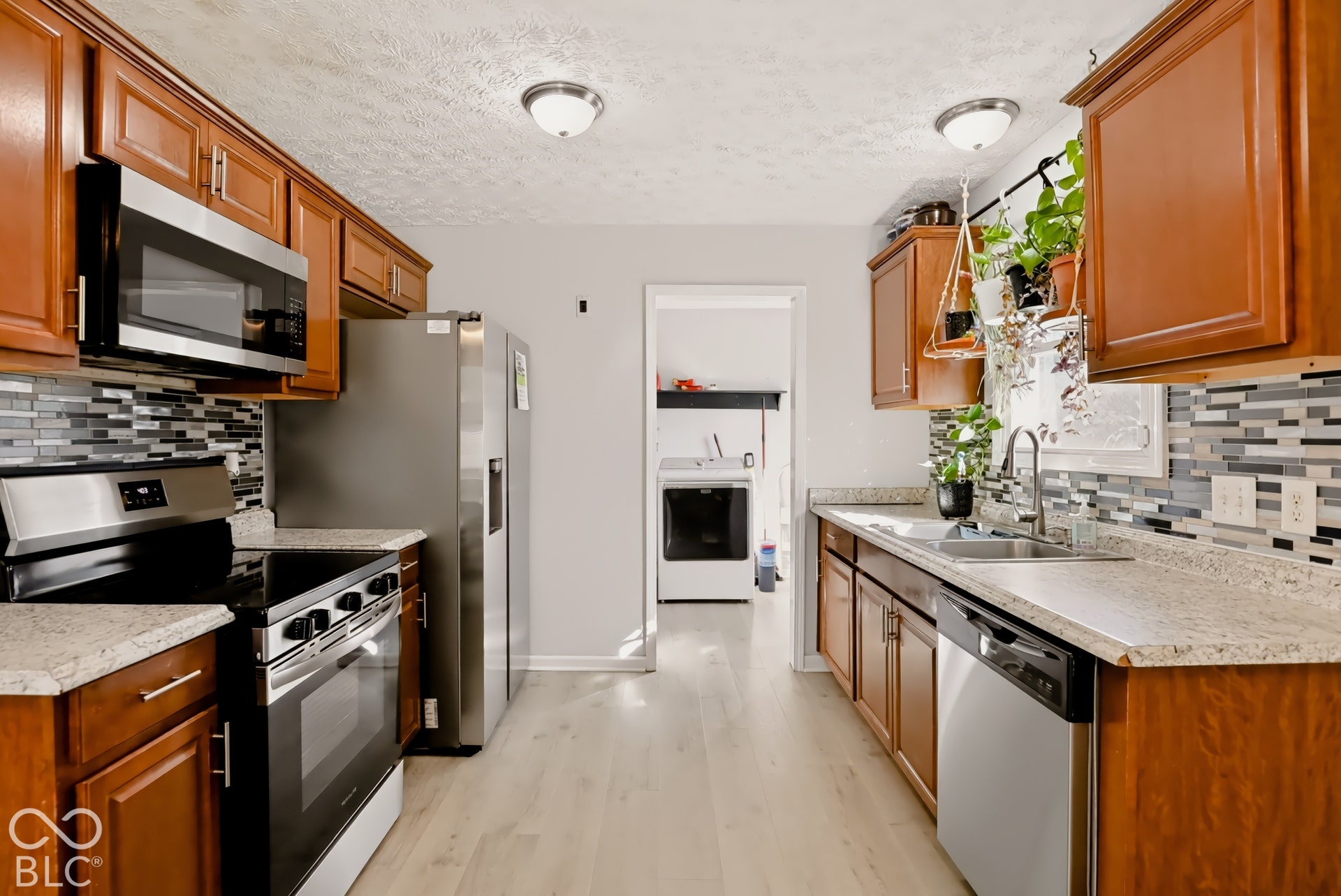 Kitchen featuring natural-finish cabinetry, stone-patterned countertops, a stainless steel appliance package, and a multi-tonal linear tile backsplash with wood-finish flooring
