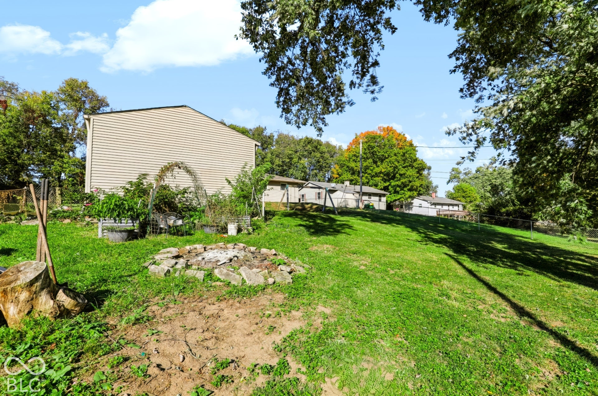 Expansive backyard featuring a stone-bordered fire pit, metal garden trellis, mature trees, and chain-link perimeter fencing