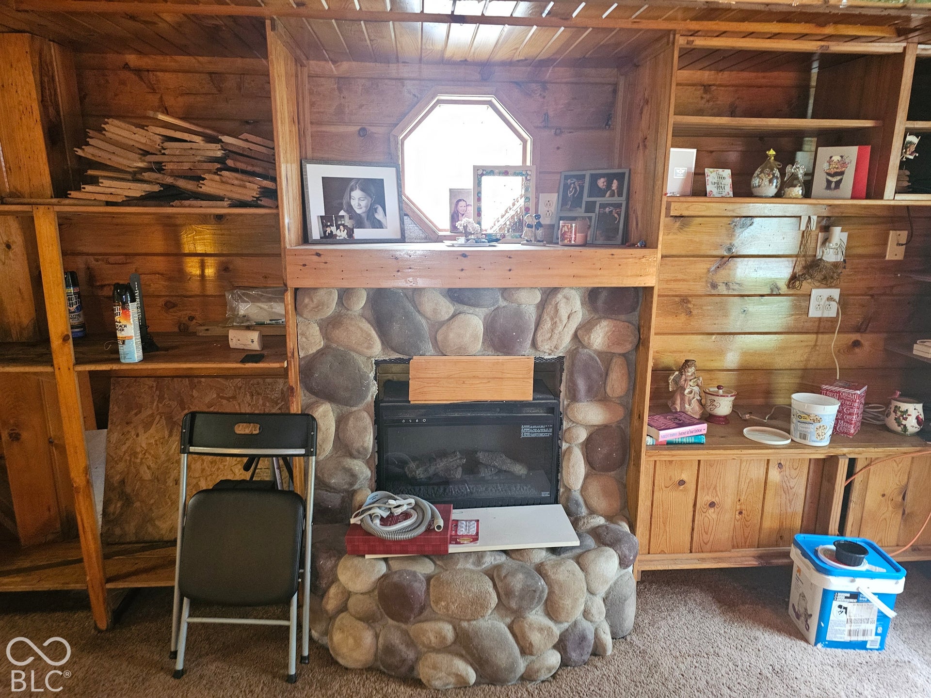 Wood-paneled walls and ceiling, stone-surround fireplace, built-in shelving, and octagonal accent window