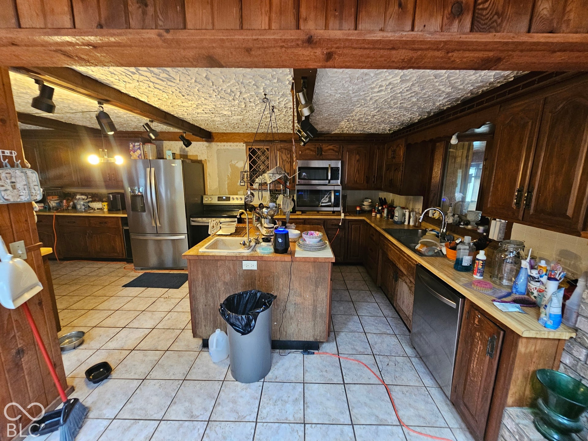 Kitchen featuring wood cabinetry, central island with an integrated sink, stainless steel appliances, ceramic tile flooring, and exposed ceiling beams