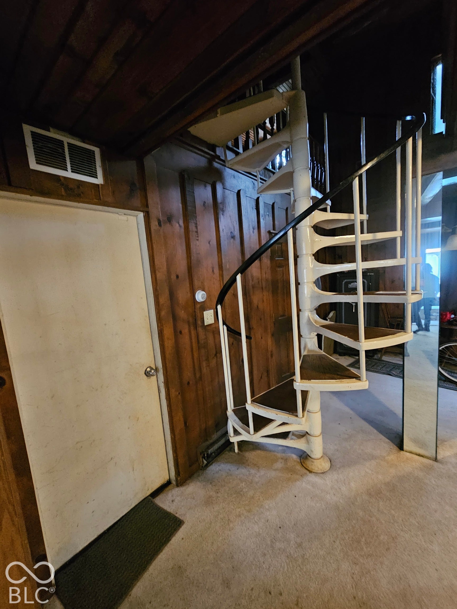 White spiral staircase featuring metal construction with a black handrail and wood-finished treads, set against dark wood paneling