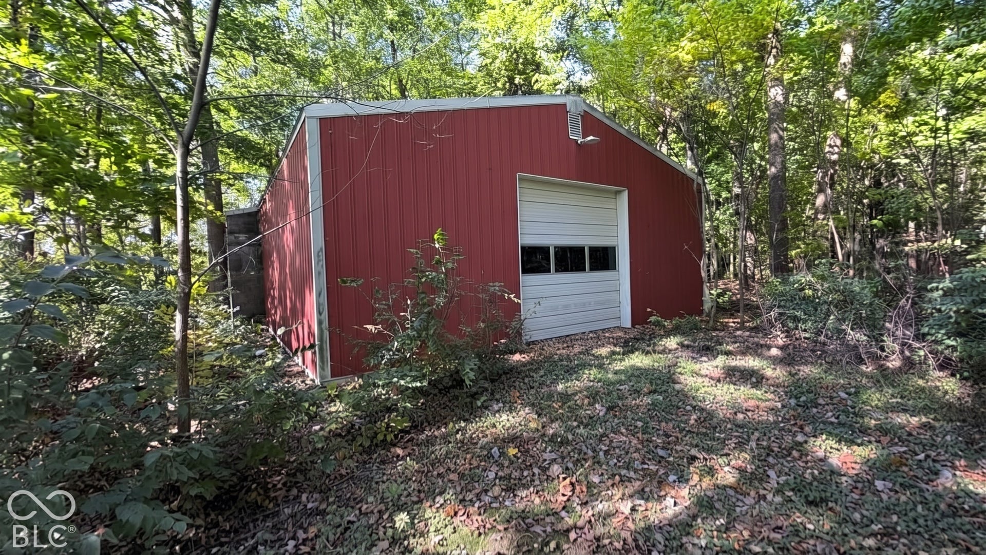 Detached metal outbuilding featuring a vertical ribbed exterior, a roll-up garage door with integrated glazing, and an exterior light fixture, situated on a wooded lot