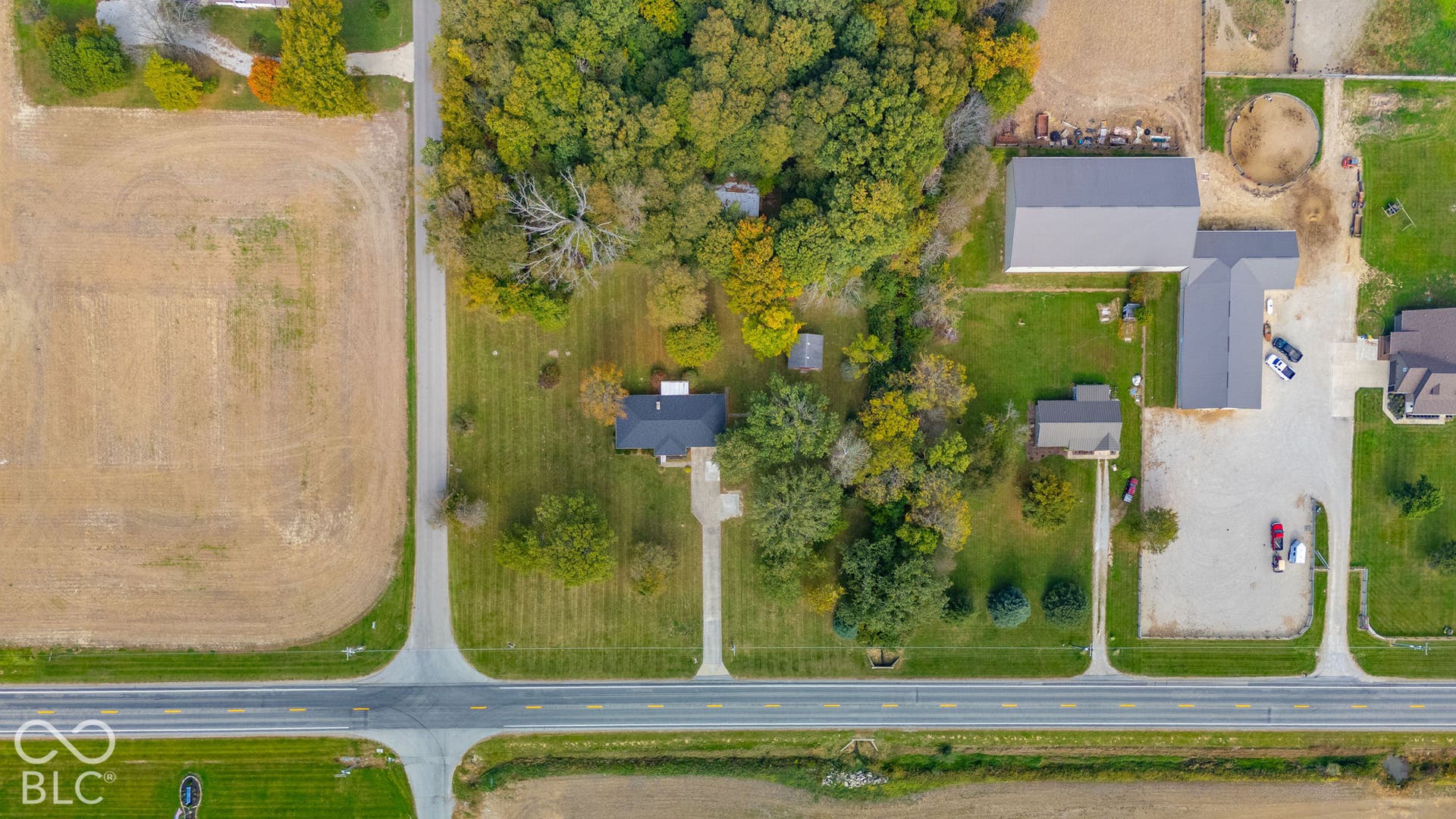 Aerial view featuring extensive acreage, multiple structures, mature trees, open lawn space, and paved driveway access