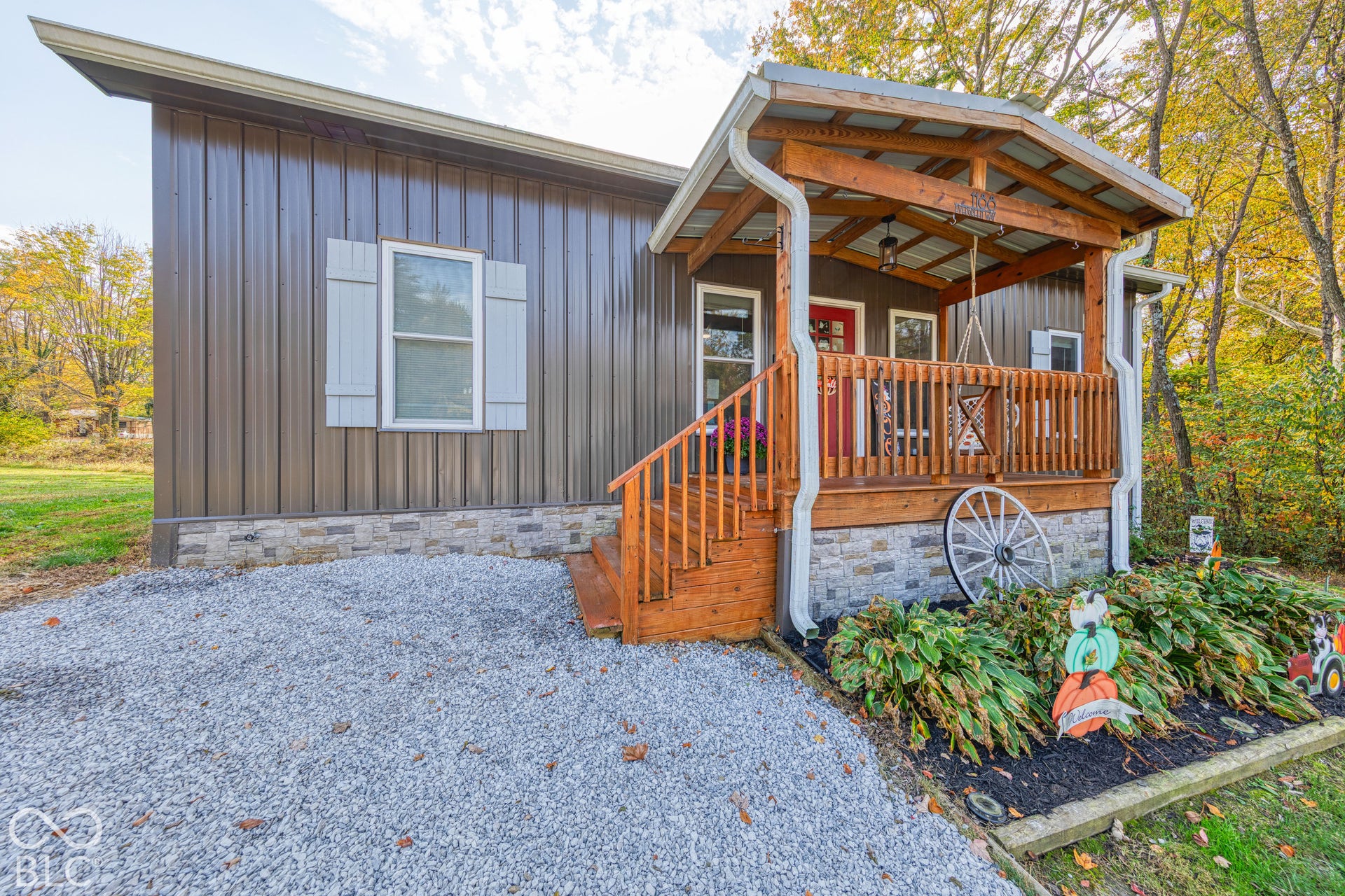 Metal-sided exterior with stone veneer skirting, covered front porch featuring a wooden railing, and a gravel driveway