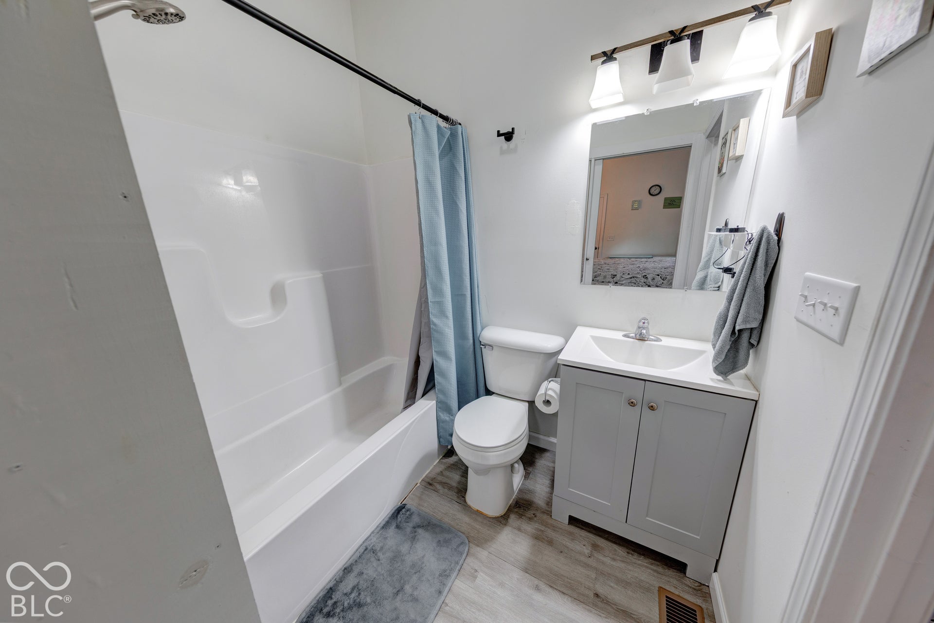 Bathroom featuring a vanity cabinet with a stone-top integrated sink, wood-finish flooring, a white bathtub and shower surround, and a three-light fixture