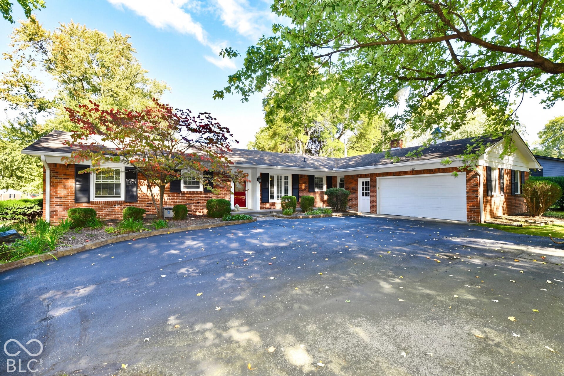 Single-story brick exterior featuring a multi-car attached garage, expansive asphalt driveway, decorative window shutters, and established mature landscaping