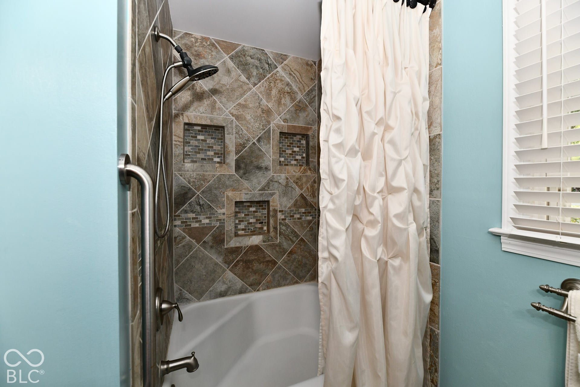 Shower featuring diagonal-set stone wall tile, built-in recessed niches, a multi-setting hand-held showerhead, metal safety grab bar, and window with adjustable blinds