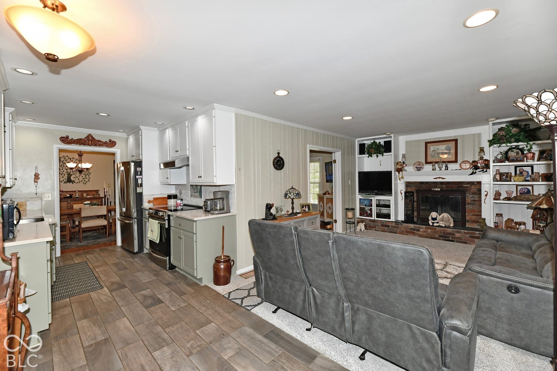 Kitchen featuring wood-finish tile flooring, white cabinetry, and stainless steel appliances, paired with a living space containing a brick fireplace and custom built-in shelving