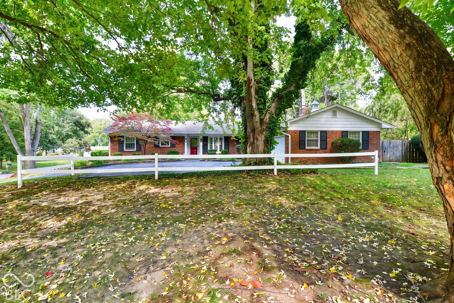 Single-story brick exterior featuring a white split-rail fence, mature trees, and shuttered windows