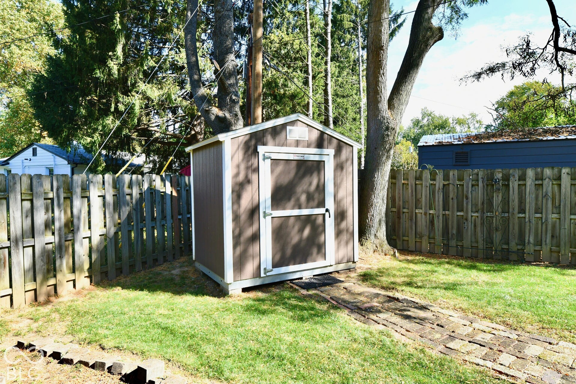 Detached storage shed with vertical panel siding, gabled roof, and white trim, situated in a fenced yard featuring a brick paver walkway