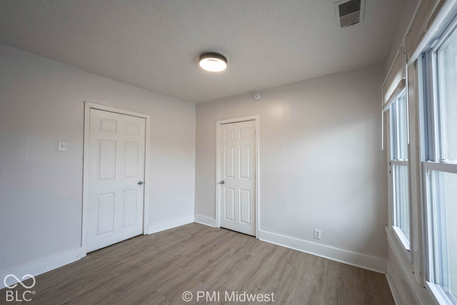 Neutral-toned interior space featuring wood-finish flooring, two six-panel doors, double-paned windows, and a flush-mount ceiling light fixture