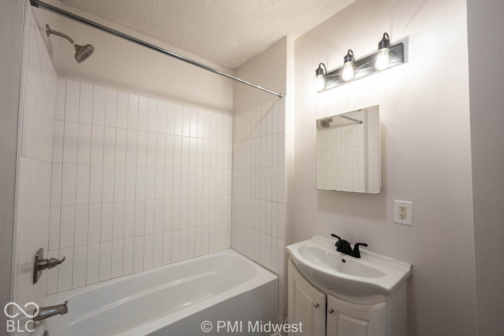 Bathroom featuring a white vanity with a ceramic basin, matte black faucet, wall-mounted medicine cabinet, white subway-style shower tile surround, and a three-light vanity fixture