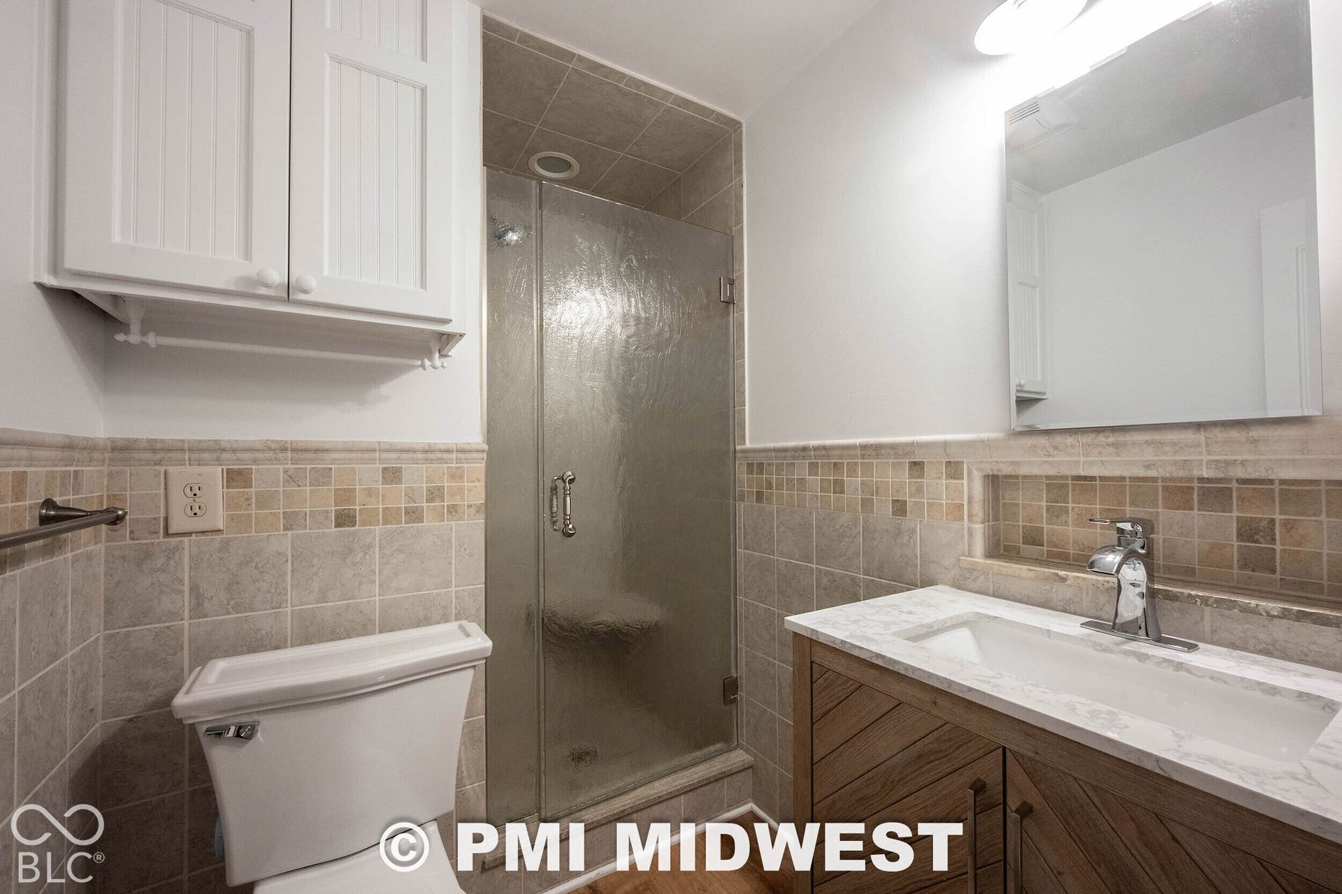 Bathroom featuring a vanity with a white countertop and rectangular undermount sink, a glass-enclosed shower with tiled walls, built-in upper cabinetry, and decorative stone tile accents