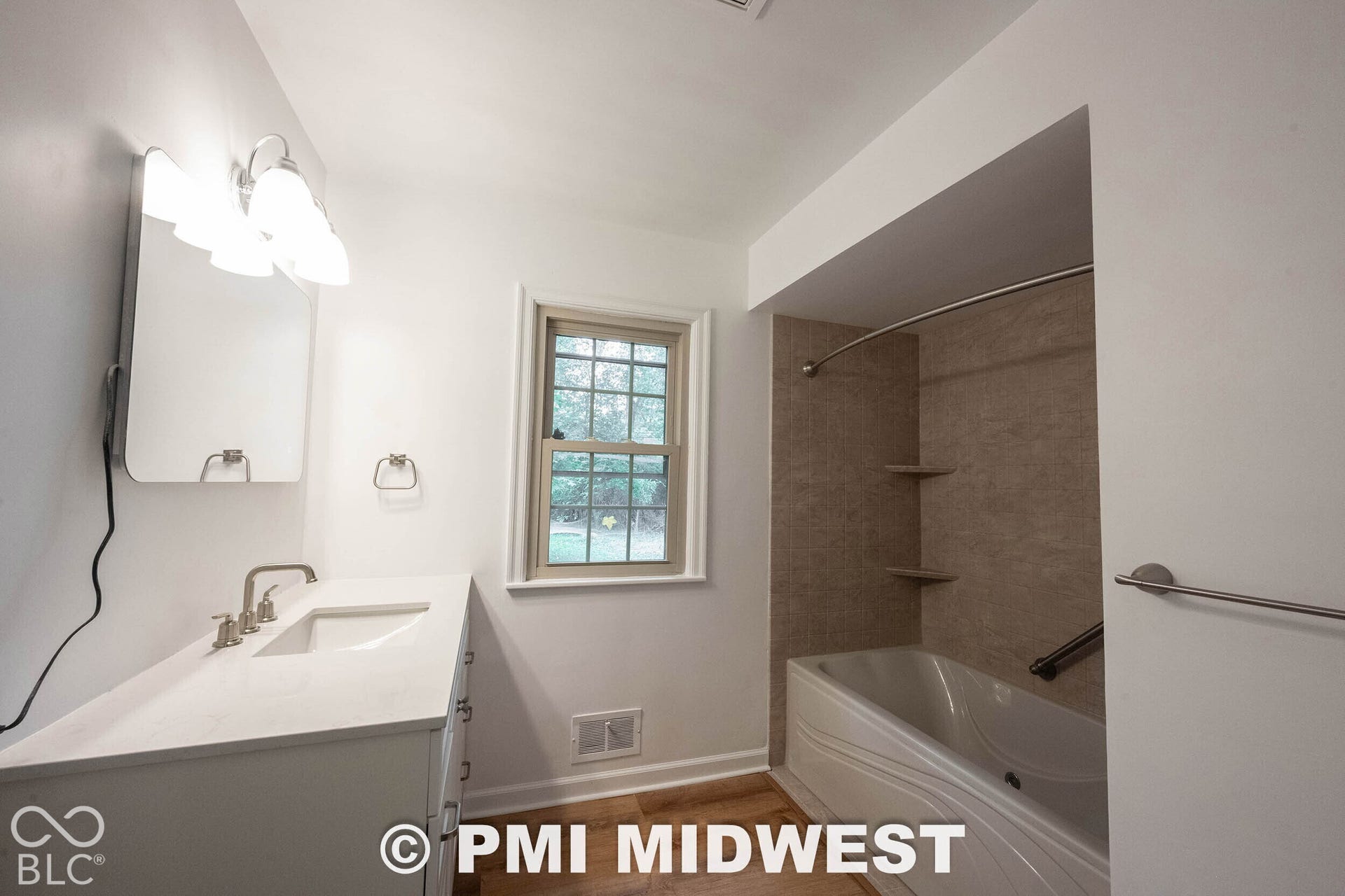 Bathroom featuring a white vanity with a rectangular basin, polished metal faucet, wall-mounted light fixture, wood-finish flooring, and a tub-shower combination with tiled surround and built-in shelving