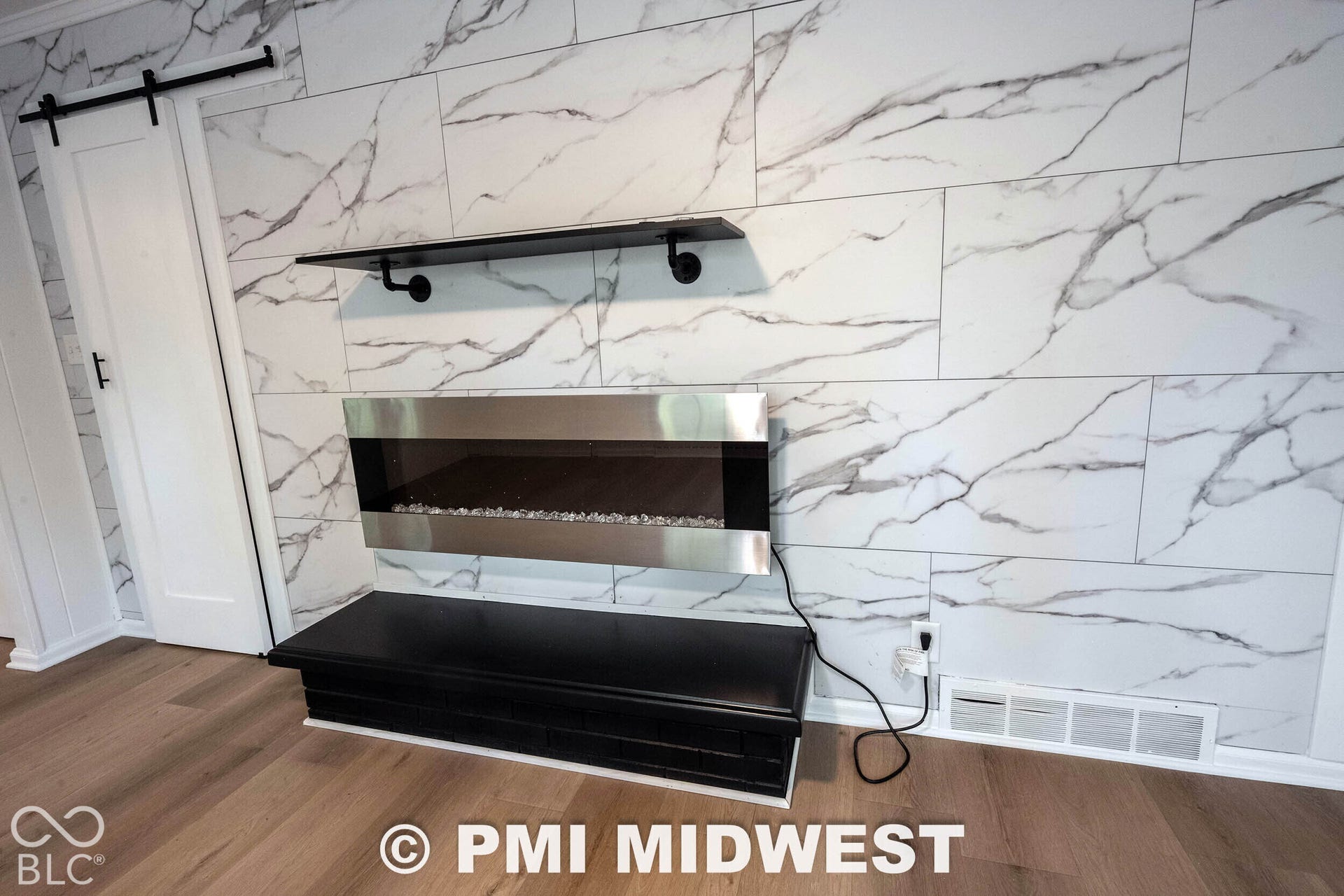 Wall-to-ceiling stone-look tile fireplace surround featuring a stainless steel wall-mounted electric fireplace, a black built-in hearth, a floating matte black shelf, and a sliding barn-style door