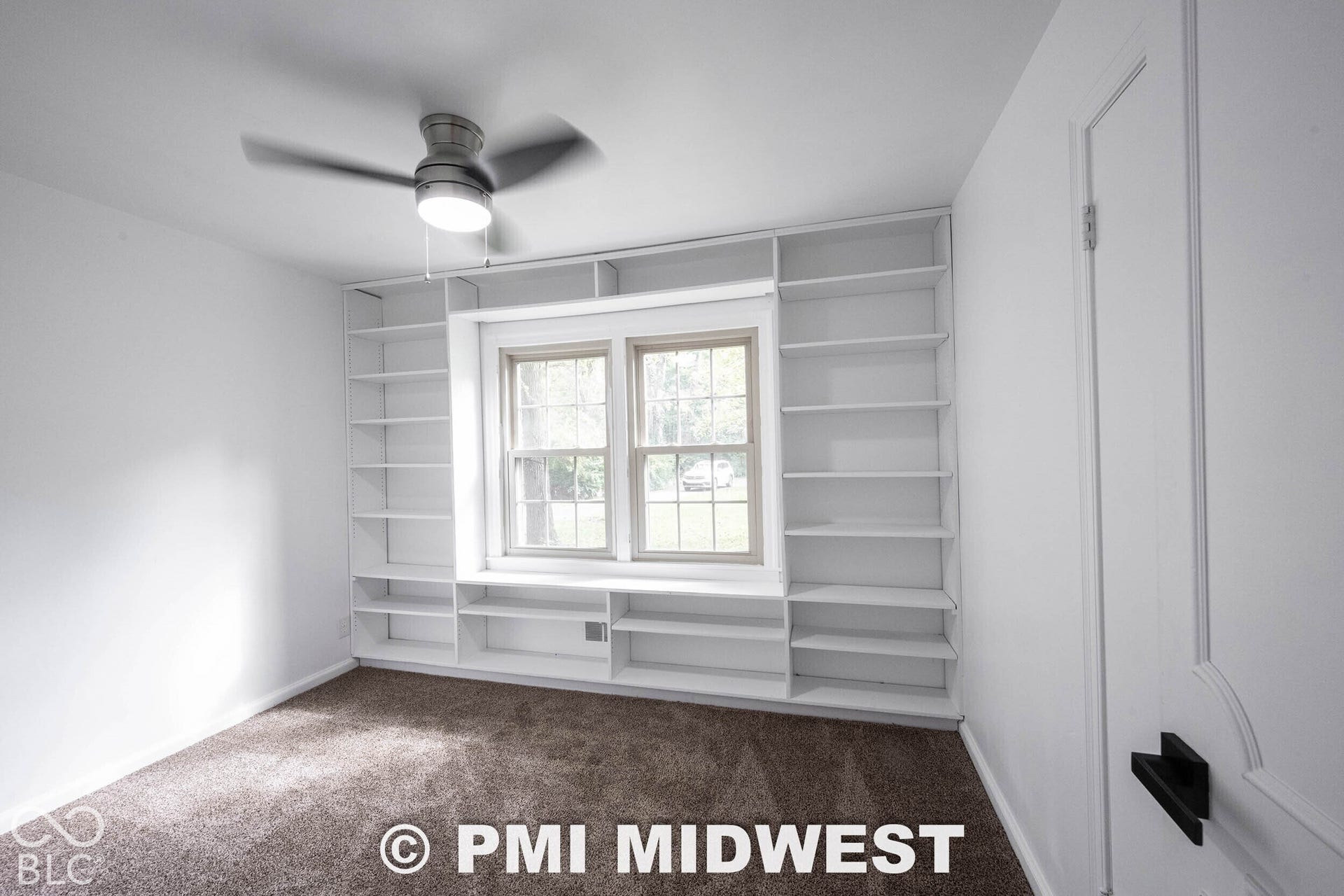 Custom floor-to-ceiling built-in shelving, double-pane windows, neutral carpeting, ceiling fan with integrated lighting, and matte black door hardware