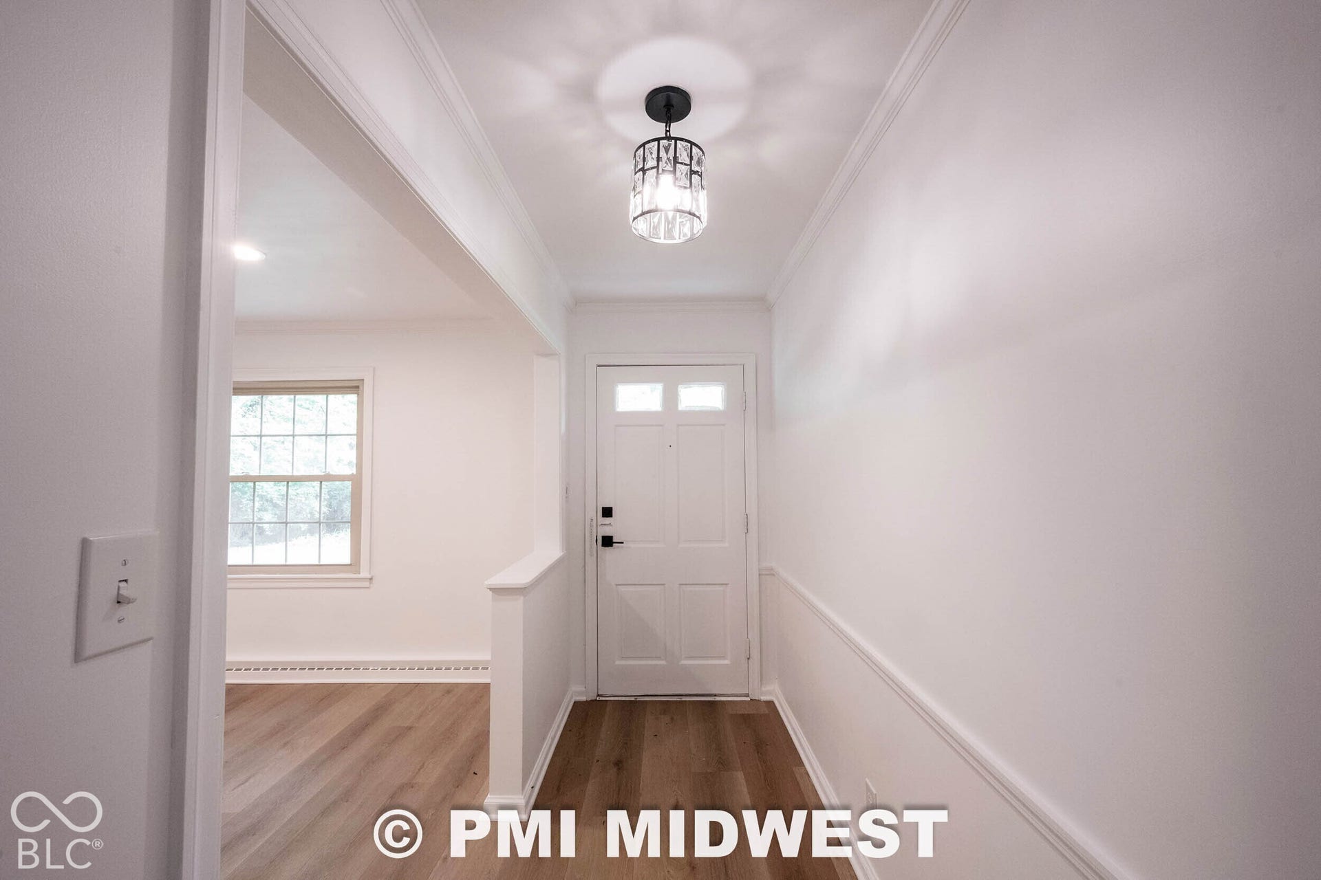 Entryway featuring wood-finish flooring, white panel door with transom window, decorative ceiling-mounted fixture, and wainscoting