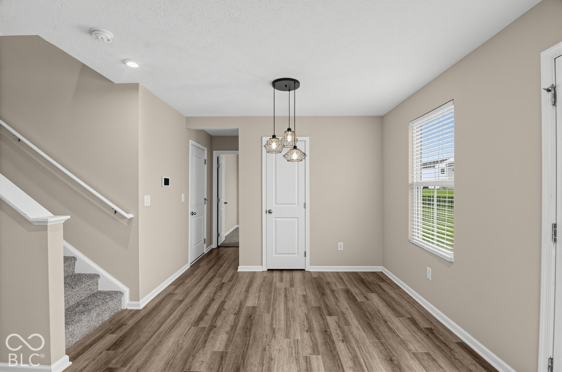 Interior space featuring wood-finish flooring, a multi-pendant light fixture, a staircase with a white handrail, neutral-toned walls, and a window with horizontal blinds
