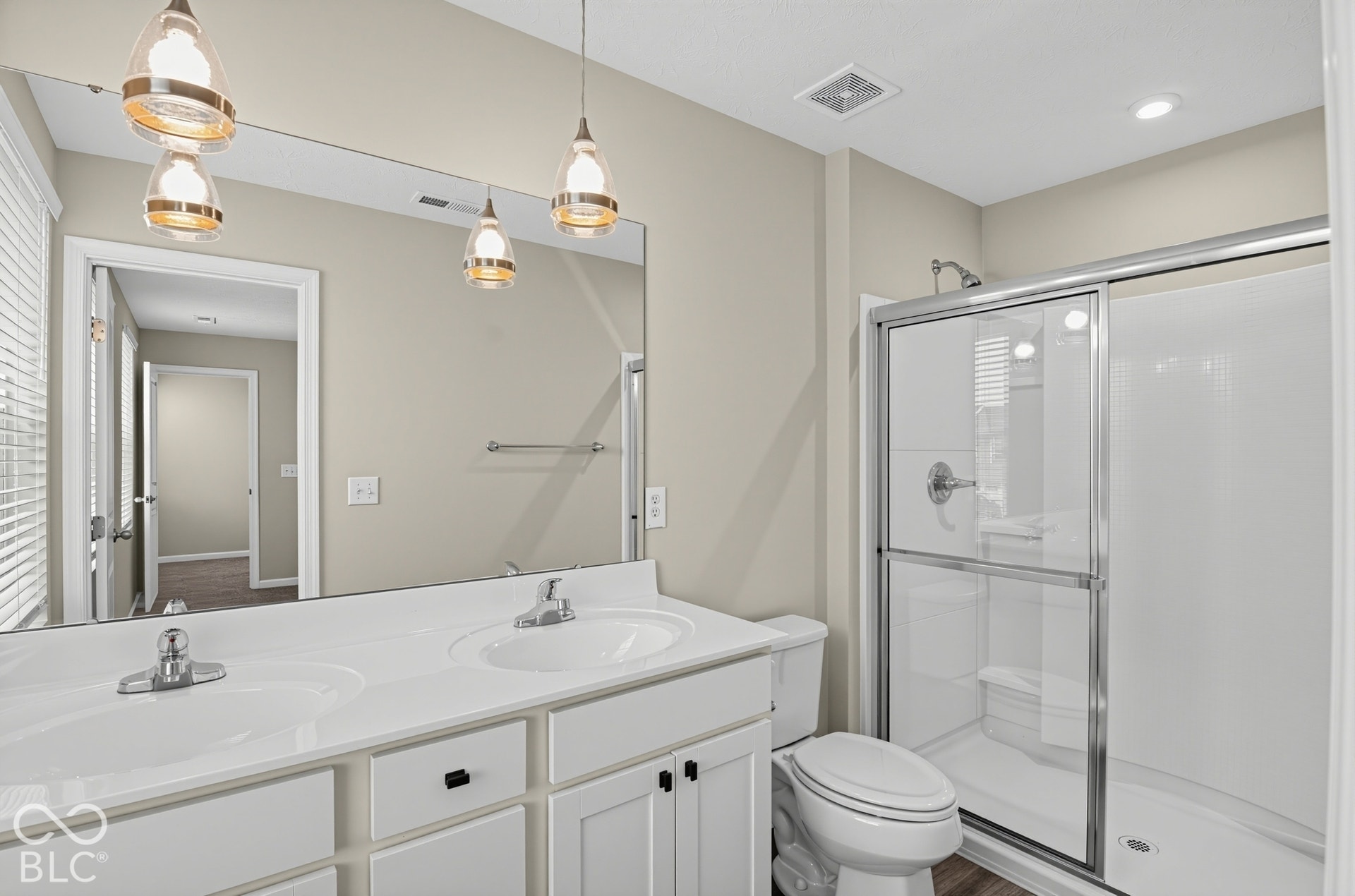 Dual-sink vanity with white cabinetry and chrome hardware, wall-to-wall mirror, pendant lighting, and a glass-enclosed shower