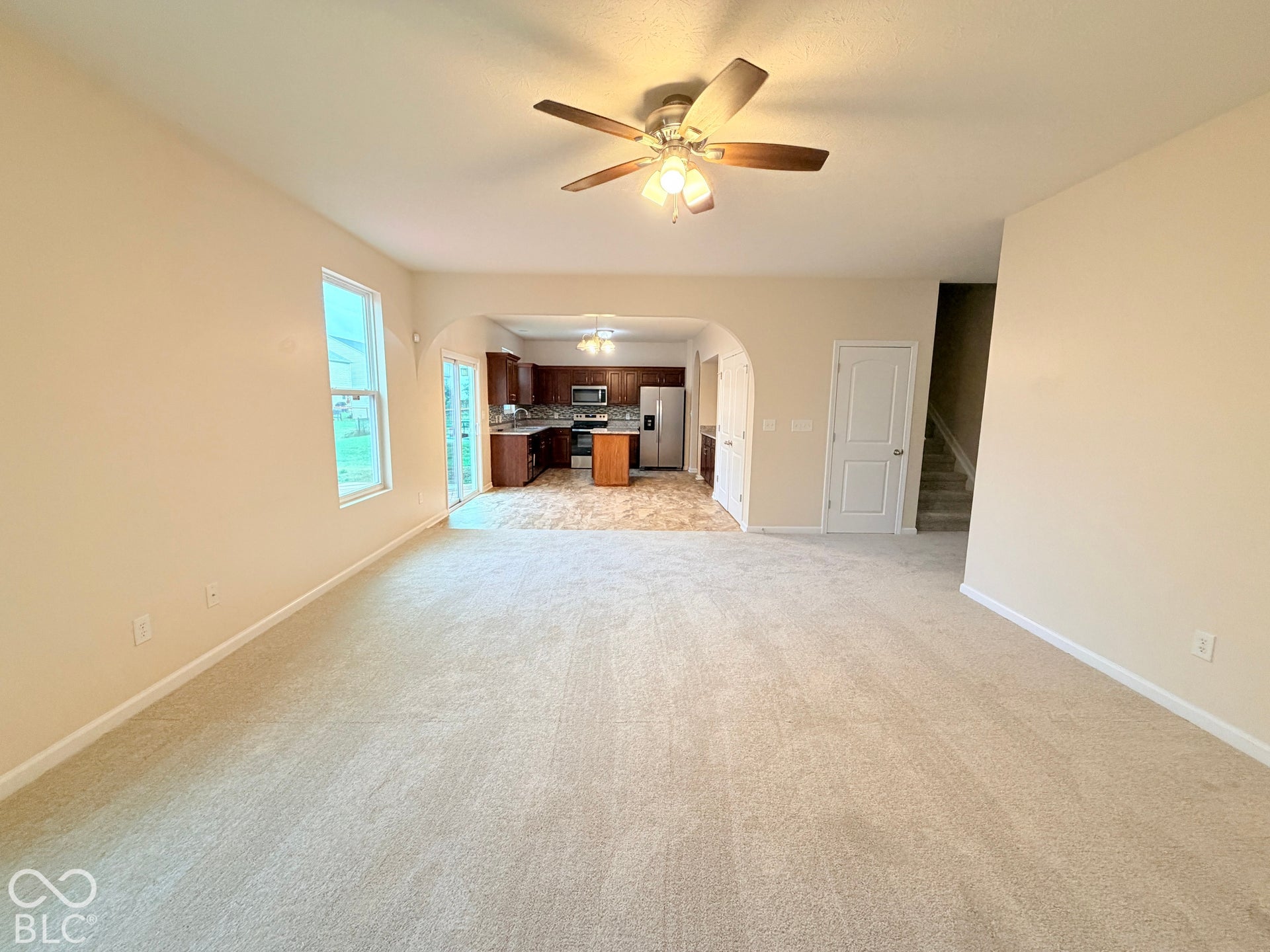 Open-concept interior featuring neutral carpeting, an arched entryway, ceiling fan with light fixture, and kitchen cabinetry with stainless steel appliances