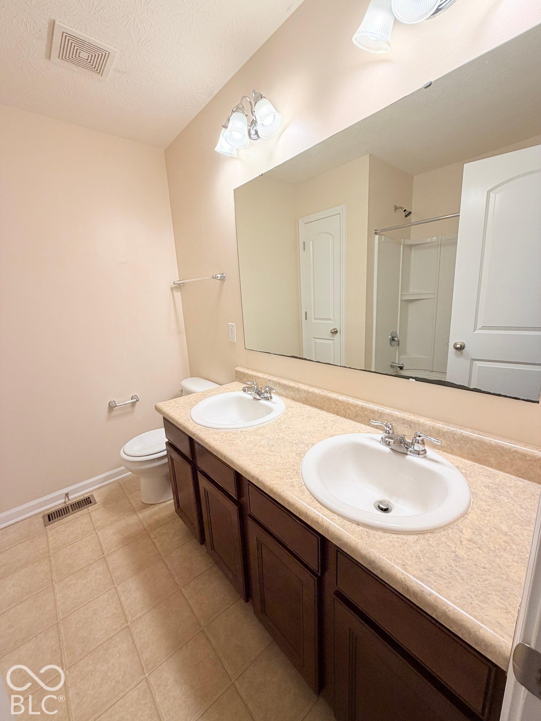 Bathroom featuring a dual-sink vanity with a solid-surface countertop, dark-finish cabinetry, a full-width wall mirror, and a shower-tub combination with an integrated surround