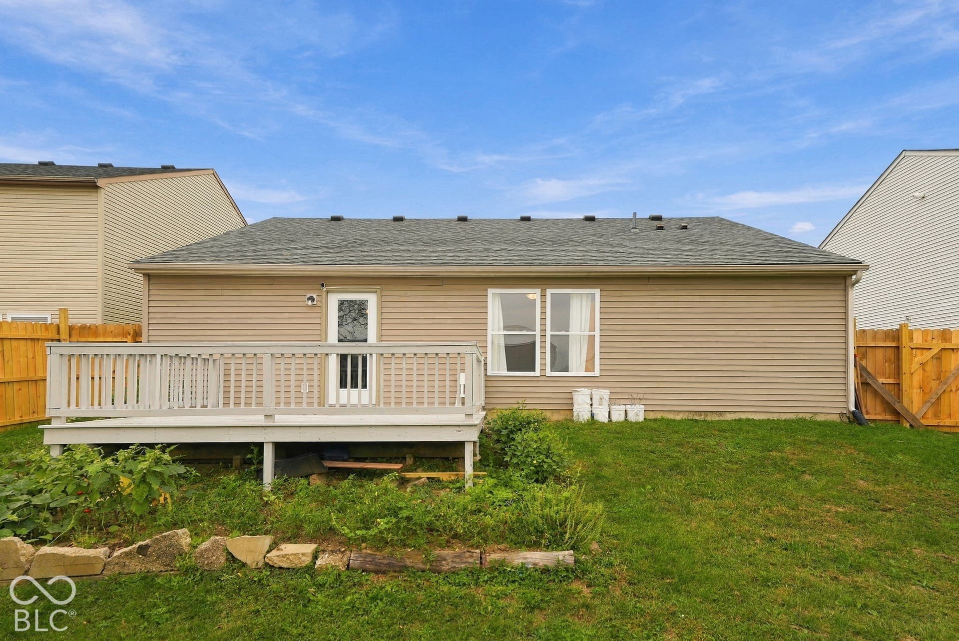 Rear exterior featuring tan vinyl siding, a raised light-gray wood-finish deck with lattice skirting, and a grass-covered backyard enclosed by wood fencing