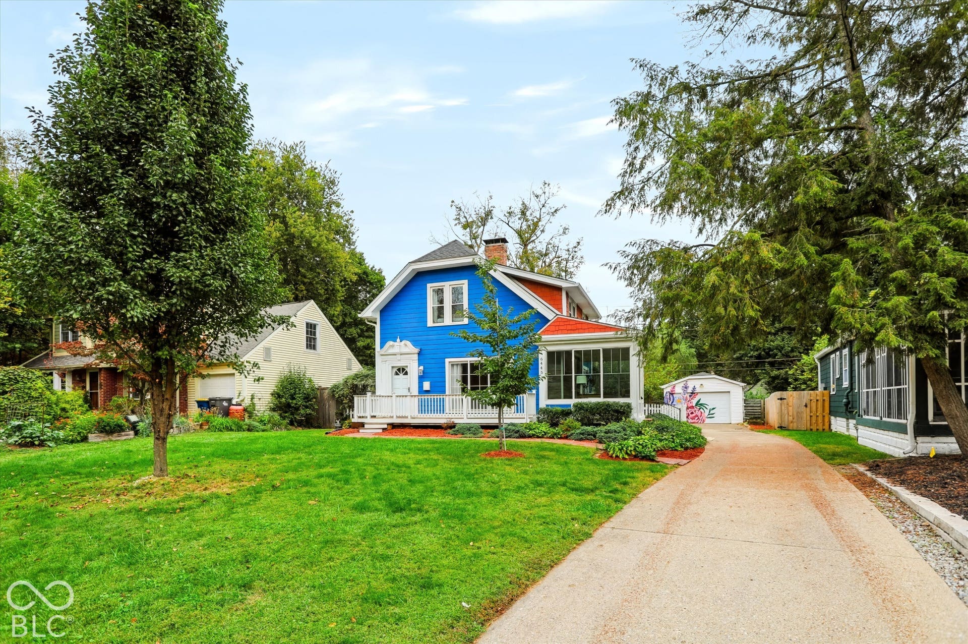 Two-story home with lap siding, a front porch with white railing, a side sunroom, a detached garage, and an expansive front lawn with mature trees