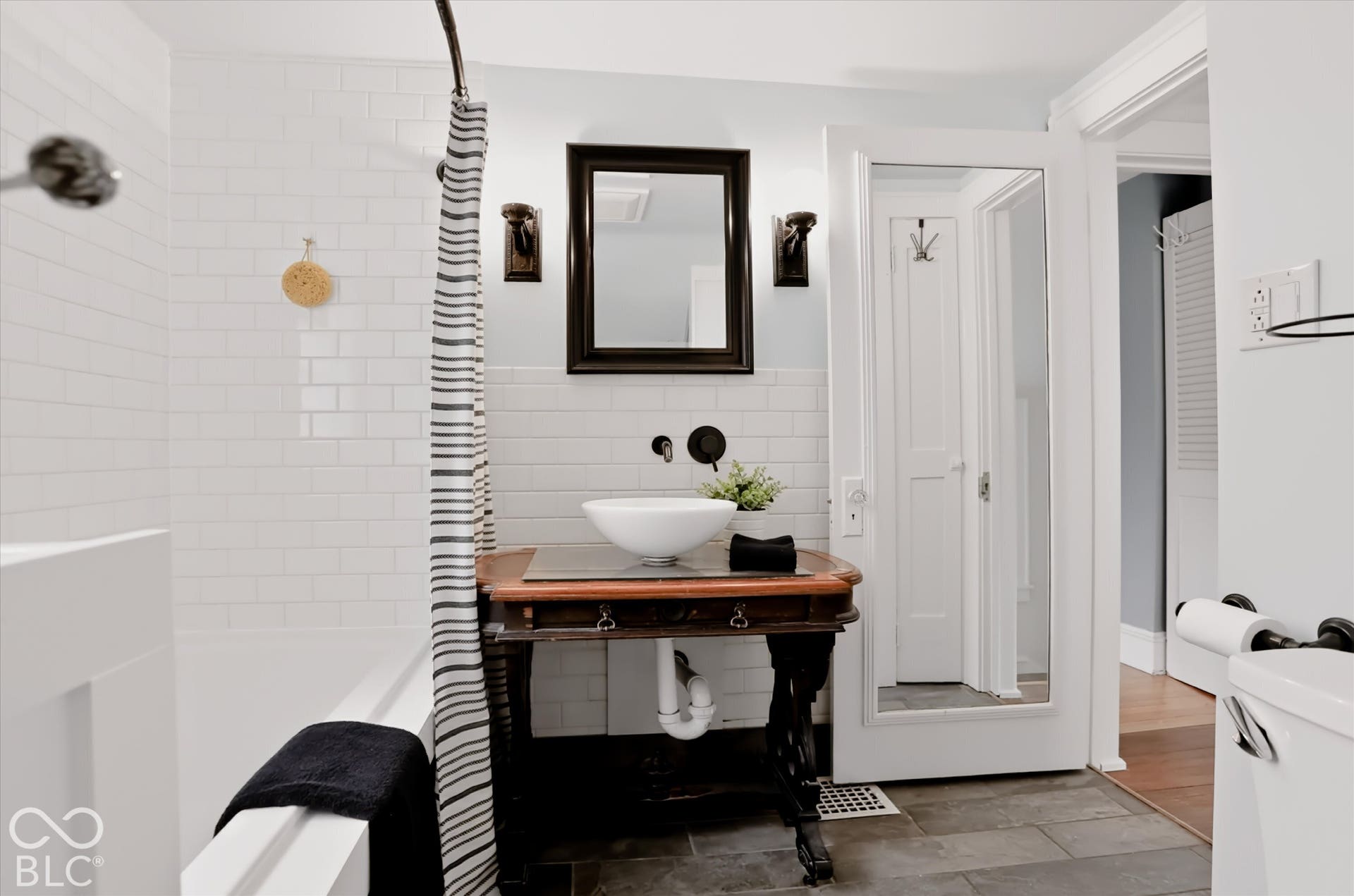 Custom vanity featuring a vessel sink with wall-mounted faucet, subway tile tub surround, mirror with dark wood frame, and stone-finish floor tile