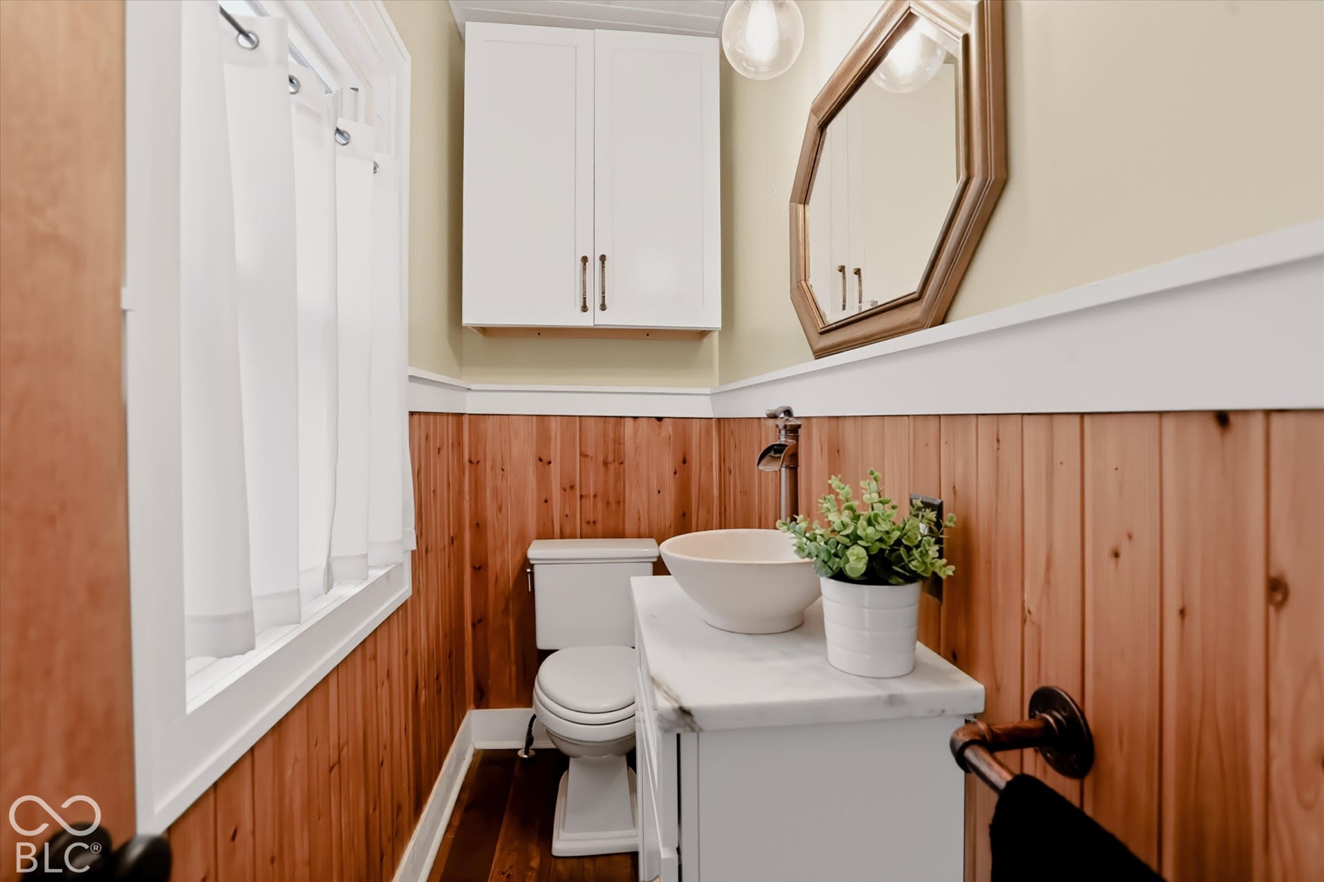Wall-mounted white cabinetry, wood paneling with horizontal trim, stone-topped vanity with vessel sink, octagonal wall mirror, and dark-finish plumbing fixtures