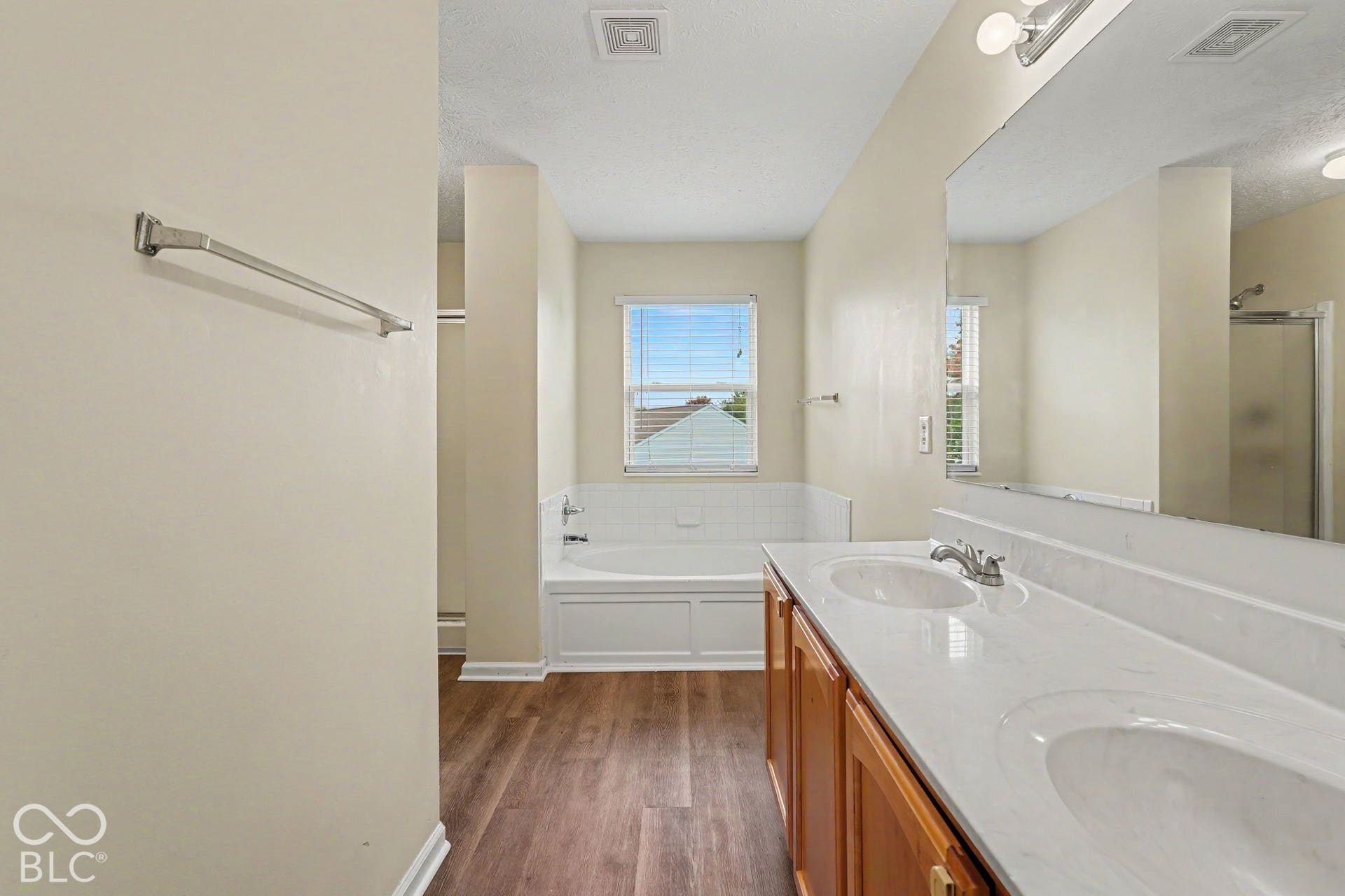 Double vanity with integrated sink basins and stone-finish countertop, soaking tub with tile surround, wood-finish flooring, and large-format wall mirror