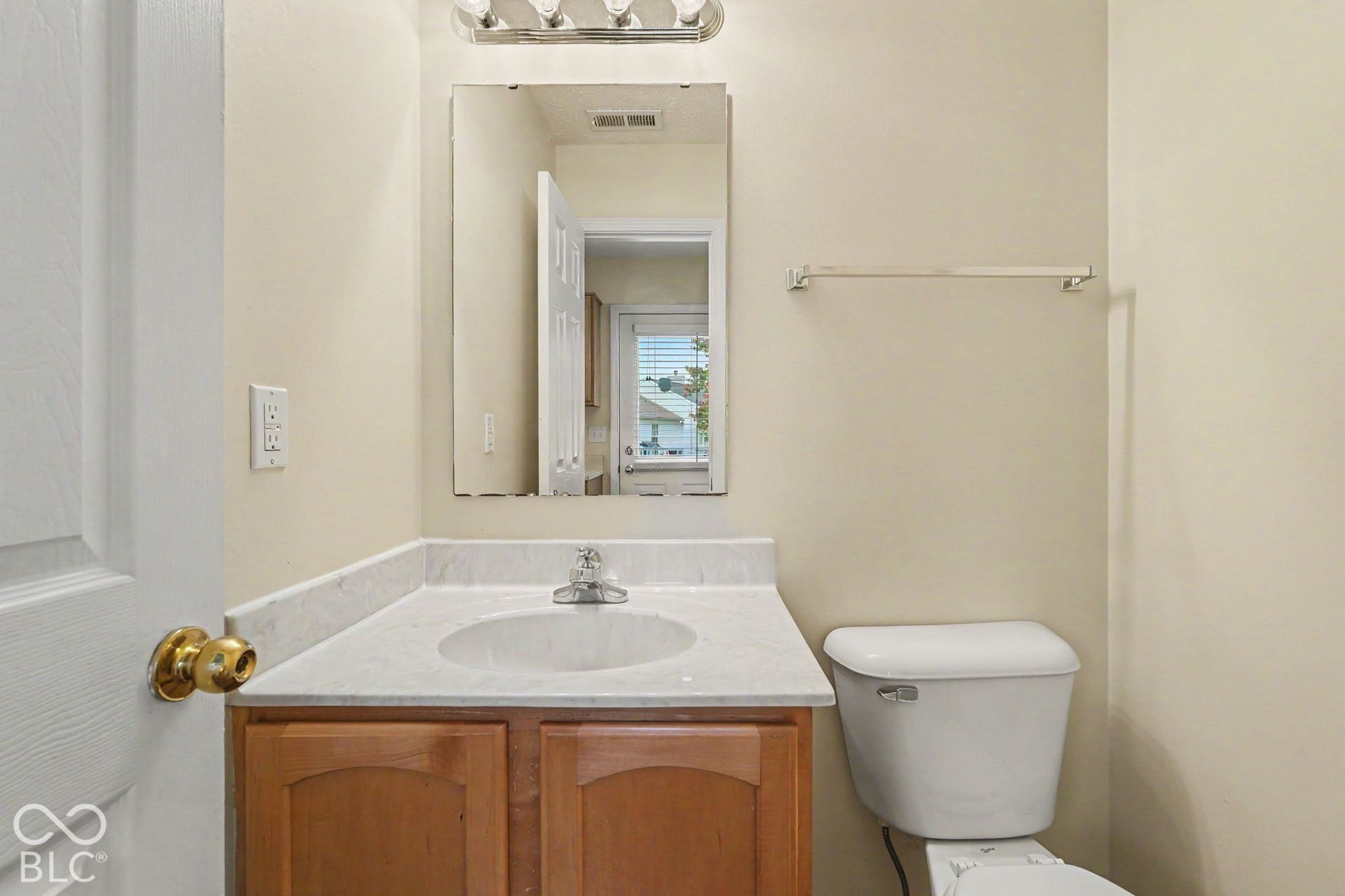 Vanity featuring a light-toned stone countertop, integrated basin, and wood-finish cabinetry, paired with a wall-mounted mirror, multi-bulb light fixture, and a white porcelain toilet