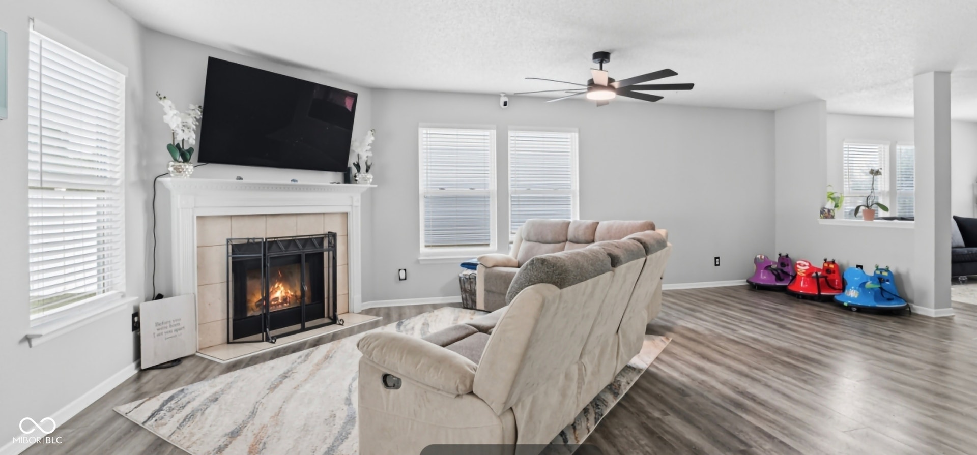 Living area featuring wood-finish flooring, a white mantel fireplace with a tile surround, multiple windows with blinds, and a ceiling fan with integrated lighting