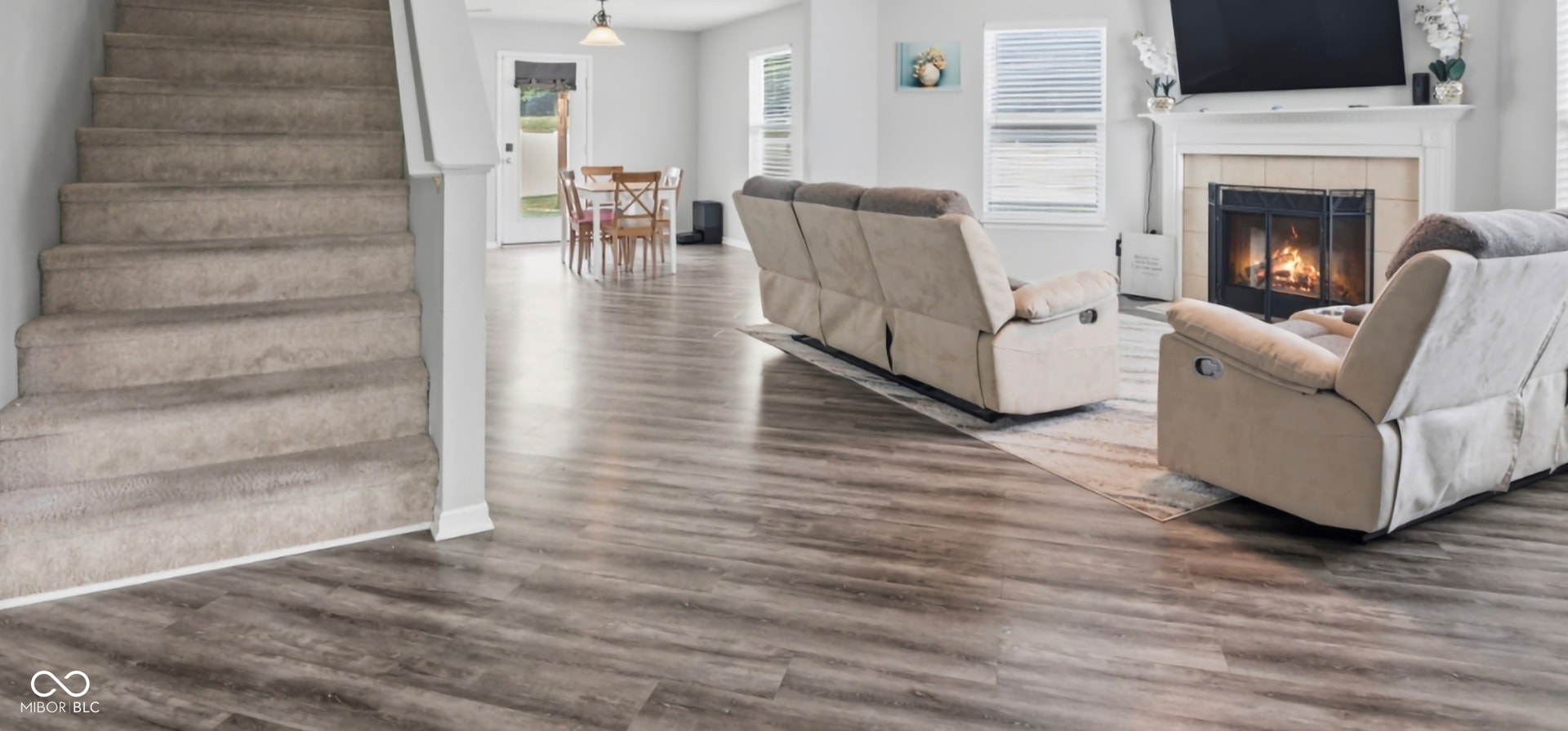 Spacious open-concept interior featuring wood-finish flooring, a carpeted staircase, a tiled fireplace with a white mantel, and white baseboards