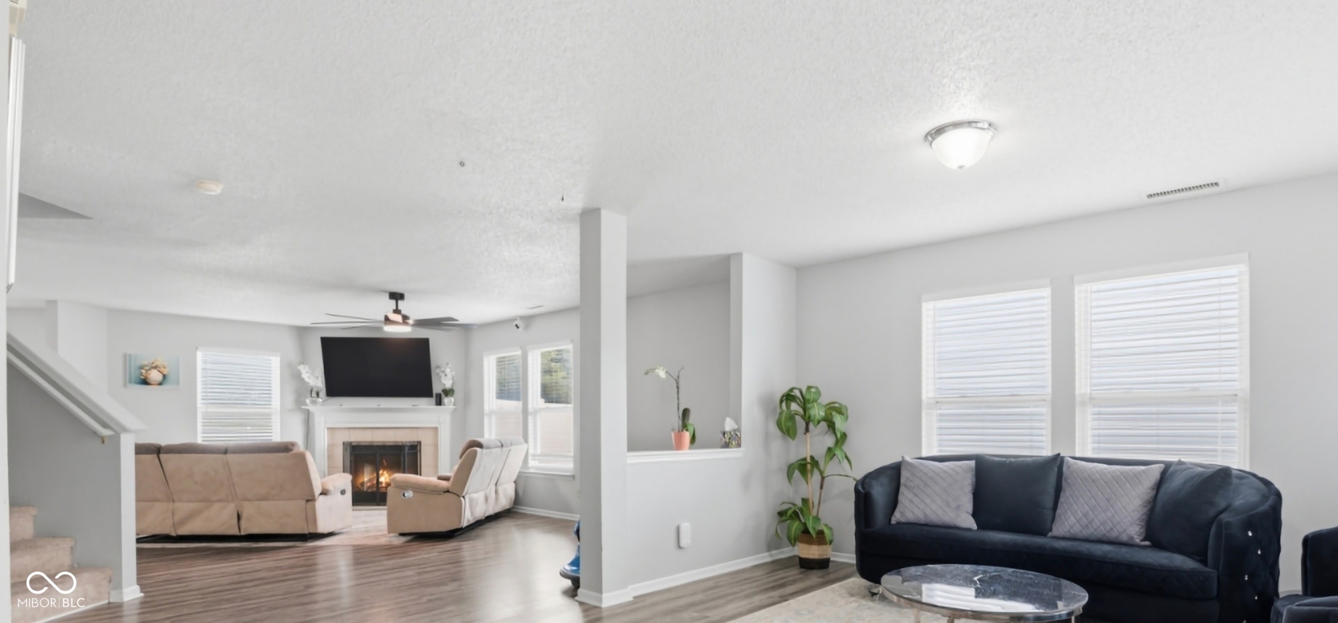 Spacious living area featuring wood-finish flooring, a fireplace with a mantel, multiple windows with blinds, and a ceiling fan