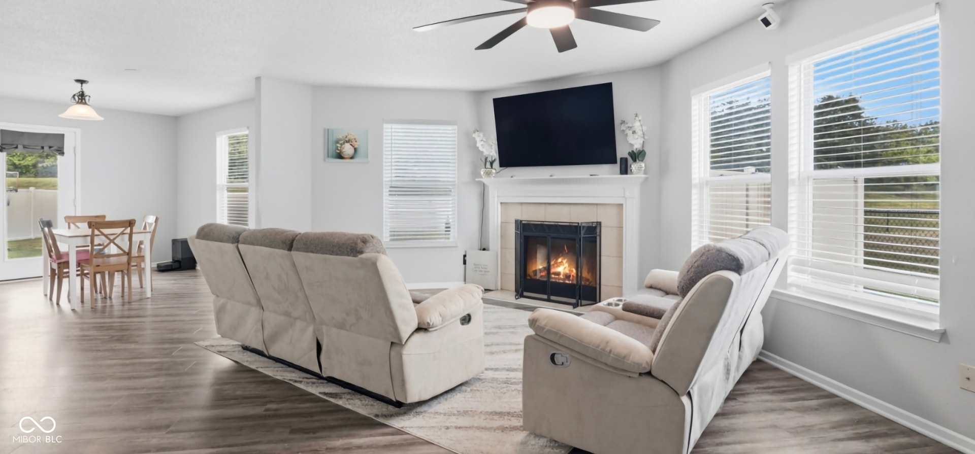 Living area featuring a fireplace with a mantel, ceiling fan with integrated lighting, wood-finish flooring, multiple windows, and a glass-paneled door