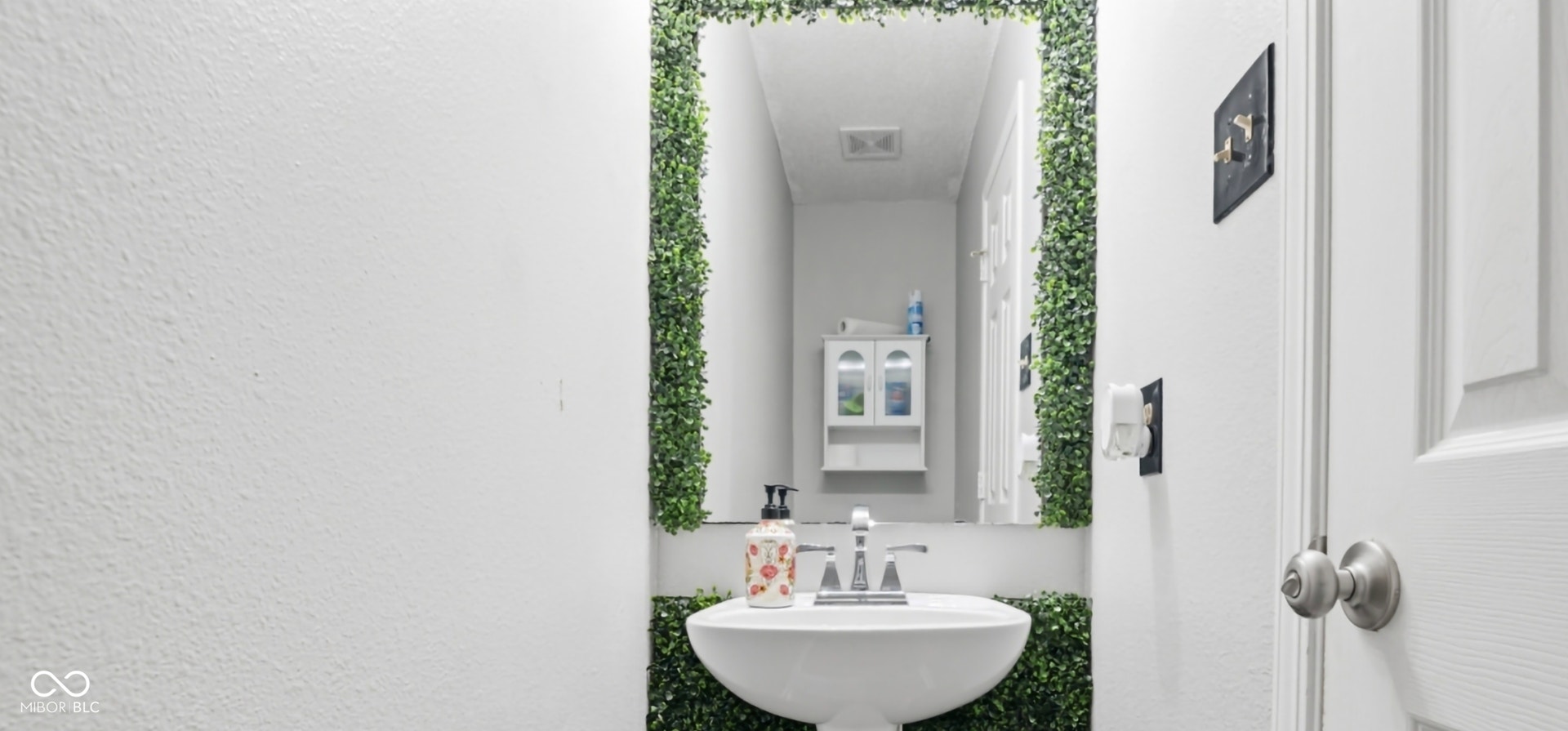 Pedestal sink with a chrome faucet, set against a mirror framed by artificial greenery