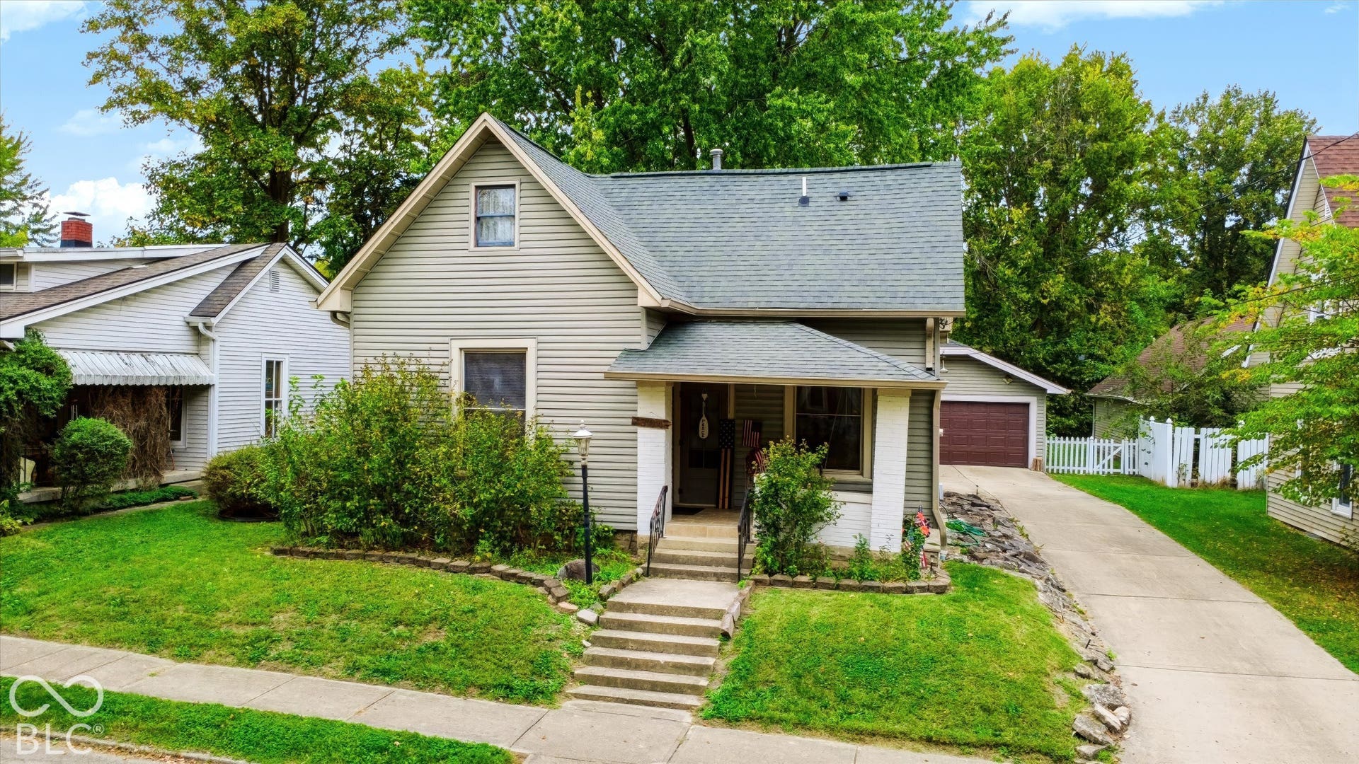 304 W 4th Street, Greenfield, IN 46140 Gable-roofed residence featuring light-toned horizontal siding, a covered front porch with squared columns, concrete steps, a private driveway, and a detached garage