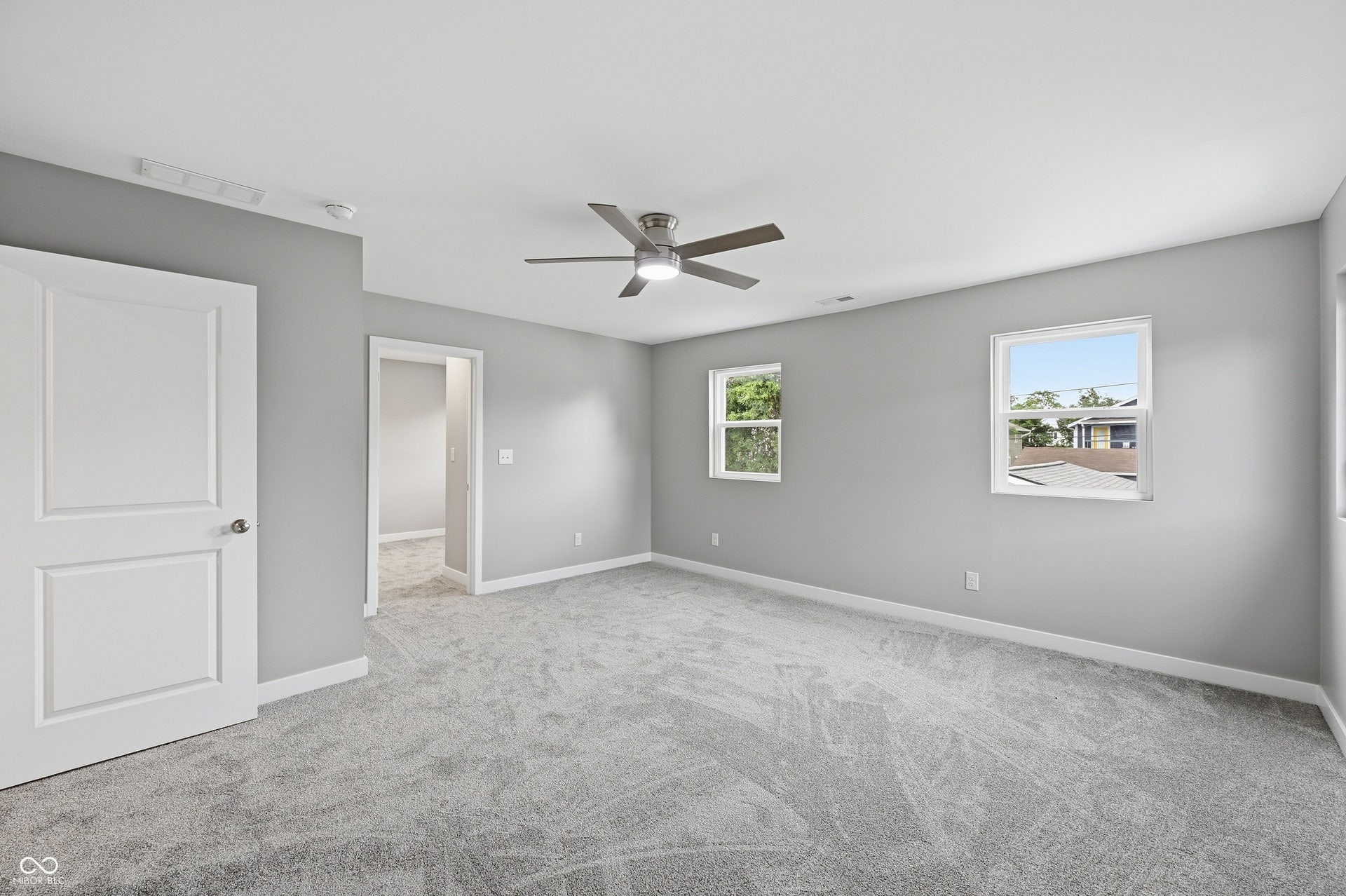 Carpeted room with light gray walls, featuring a modern ceiling fan, two windows providing natural illumination, and a white paneled door