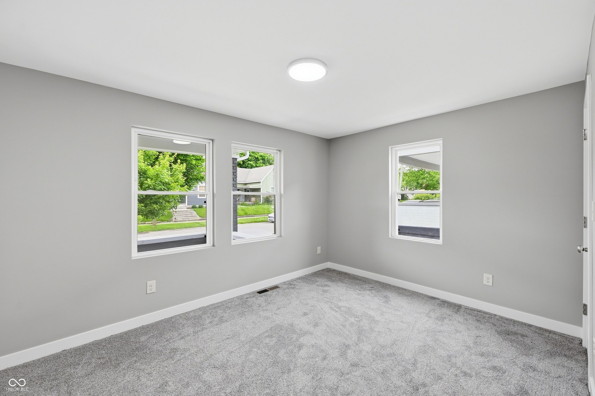 Well-lit room featuring multiple windows, neutral gray walls, plush gray carpeting, white trim, and a ceiling-mounted light fixture