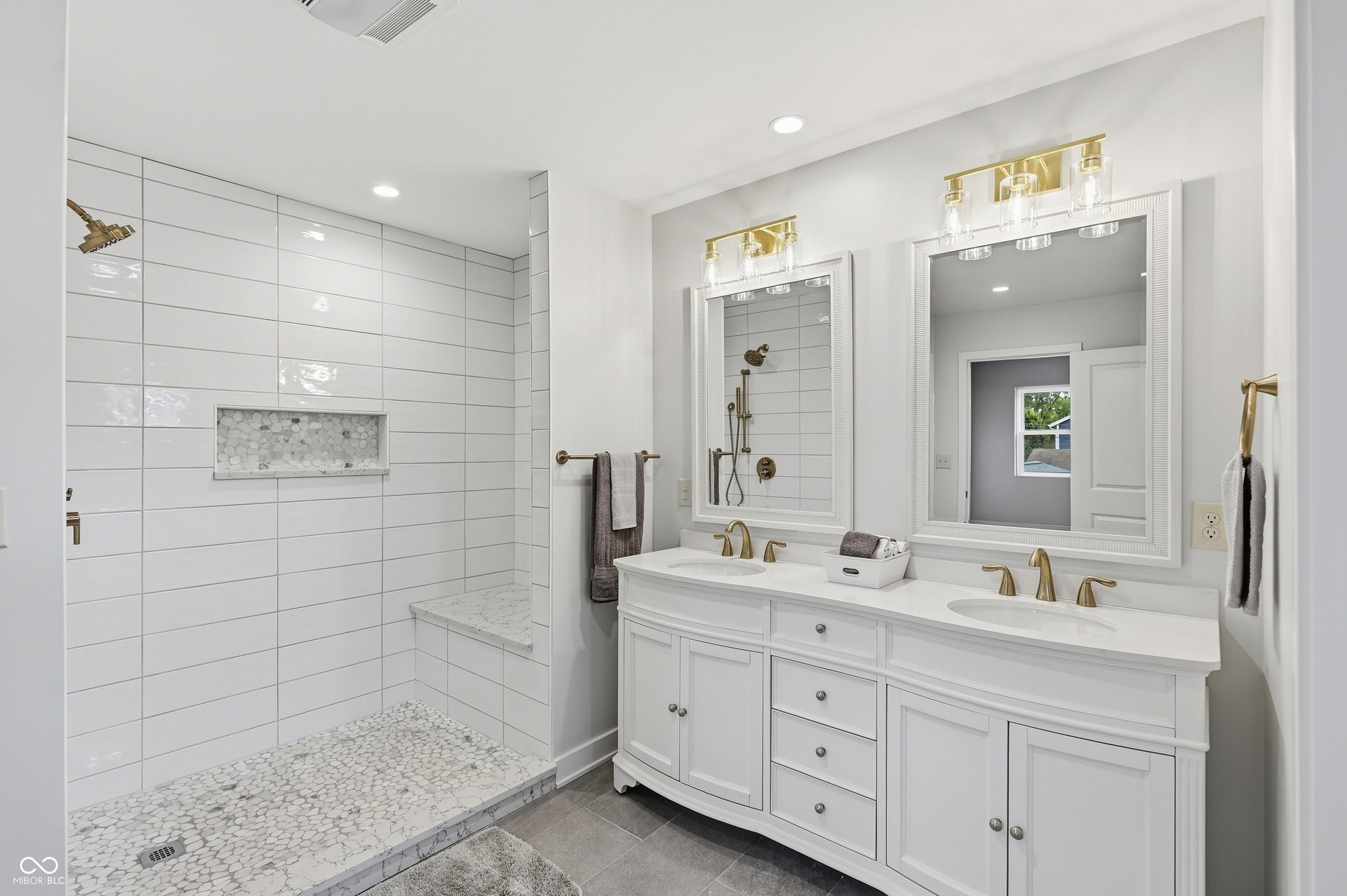 Spacious bathroom featuring a double vanity with white cabinetry and undermount sinks, bronze-finish fixtures, dual framed mirrors with overhead lighting, and a walk-in shower with white subway tile, pebble flooring, and a built-in bench