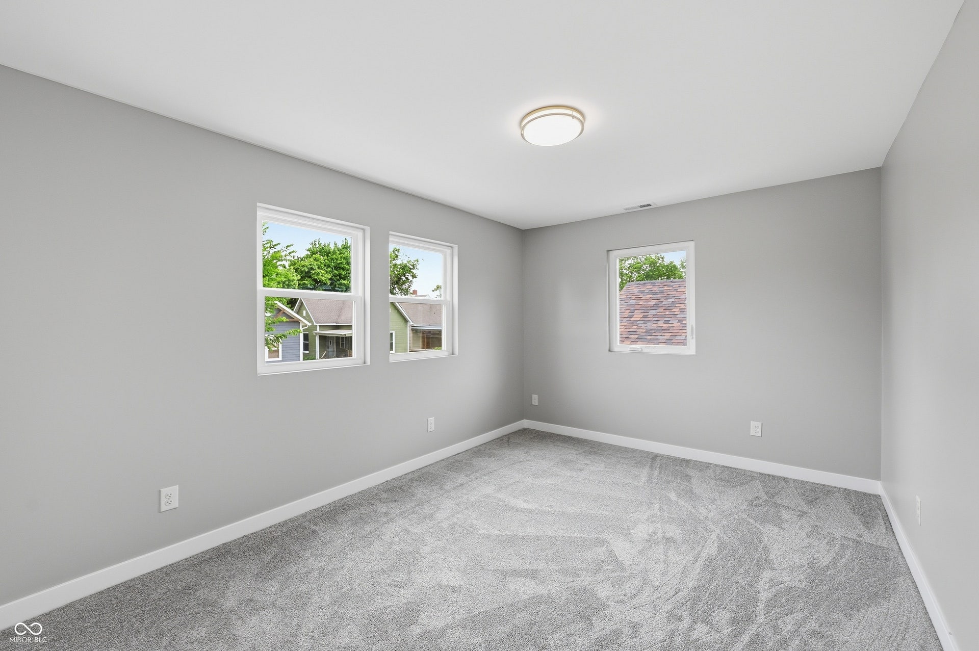Spacious room featuring light gray walls, plush gray carpeting, white trim, three windows, and a flush-mount ceiling light fixture