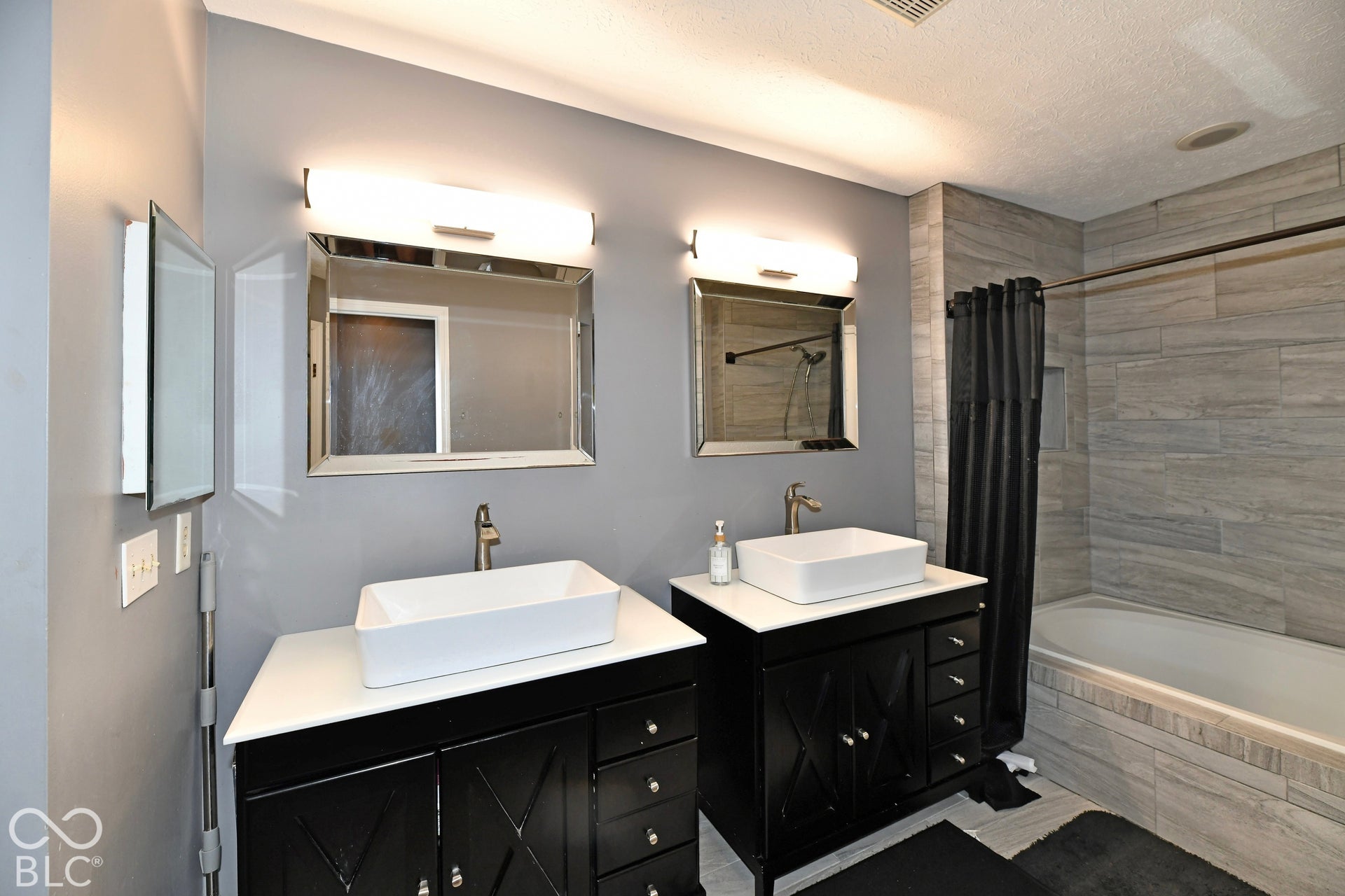 Dual vanity featuring white vessel sinks, quartz countertops, black cabinetry with X-pattern detailing, horizontal wall-mounted lighting, and a tile-surround bathtub