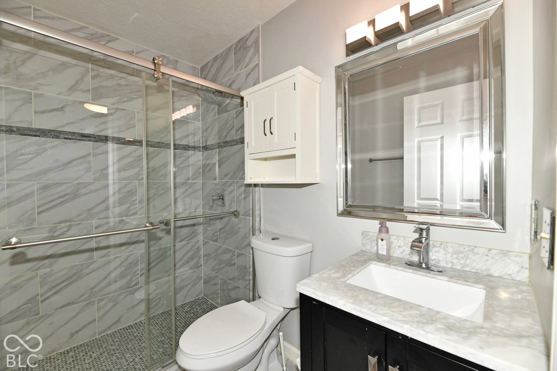 Bathroom featuring a walk-in shower with stone-finish tile surround and glass sliding doors, vanity with a light-toned countertop, undermount sink, and wall-mounted light fixture