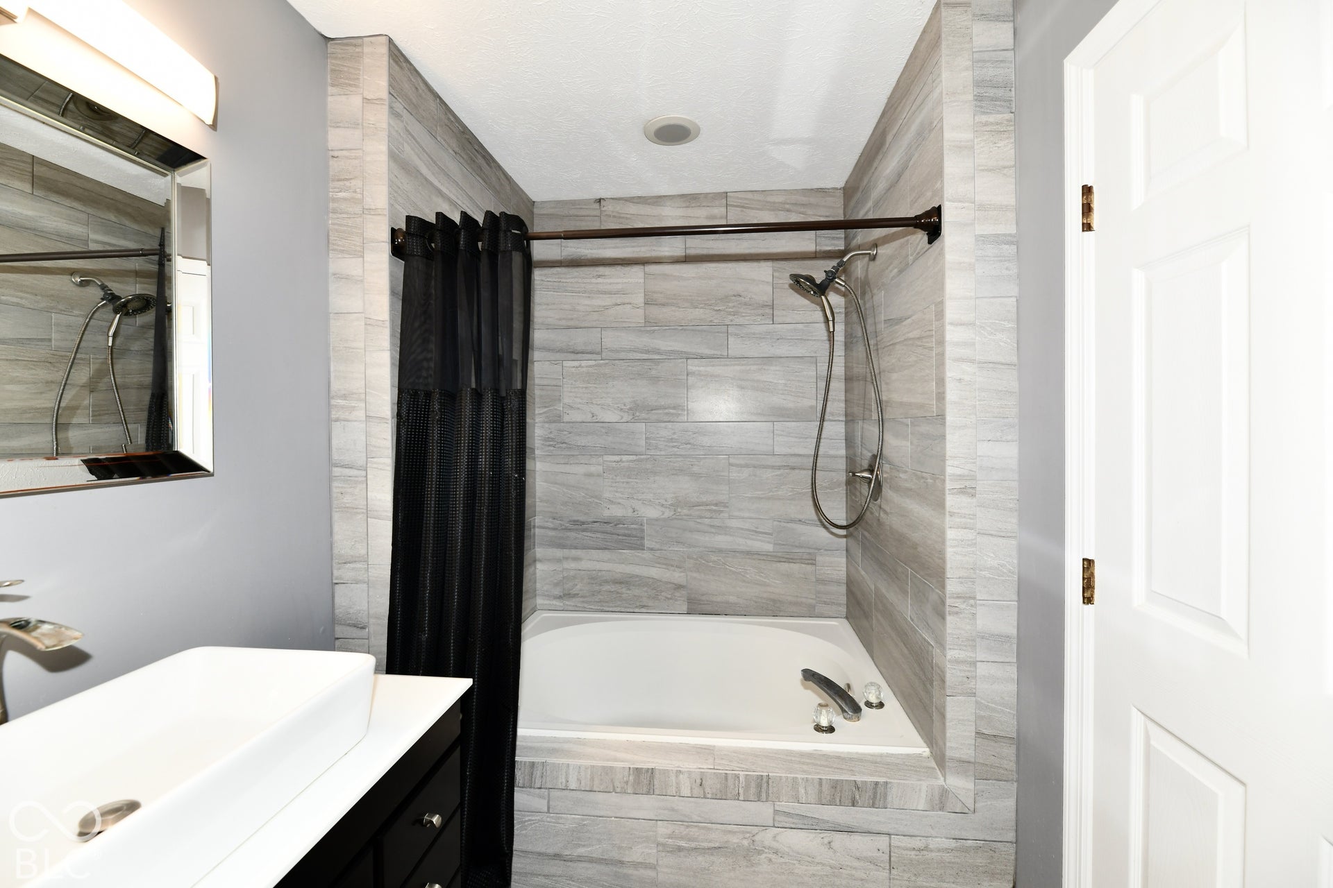 Recessed soaking tub featuring neutral-toned stone-finish tile surround, handheld shower fixture, rectangular vessel sink atop a dark vanity, and wall-mounted vanity lighting