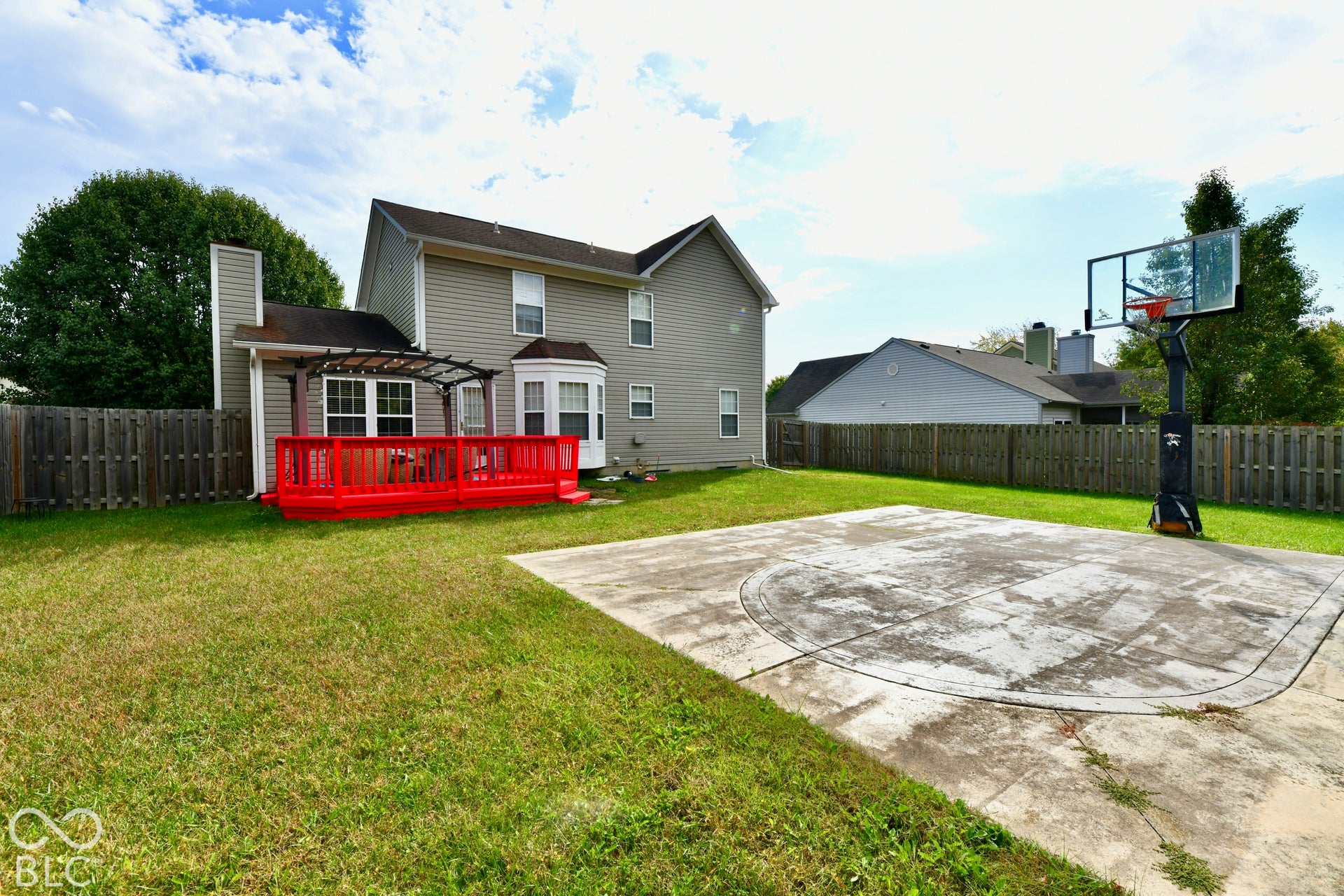 Rear exterior featuring a wood deck with an attached pergola, a paved concrete sports court, and perimeter wood fencing
