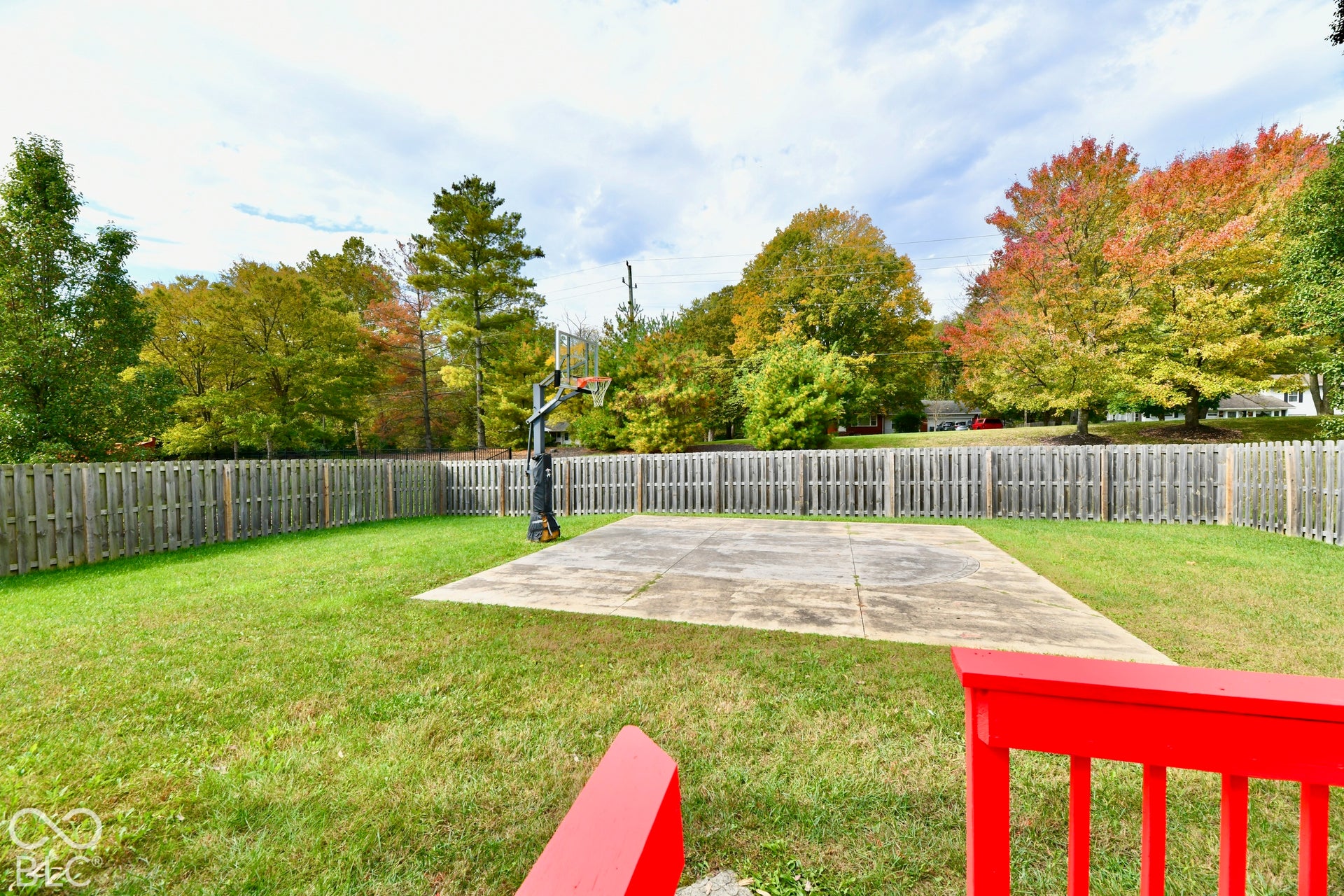 Backyard featuring a concrete court, attached basketball hoop, wood-plank fencing, and mature trees