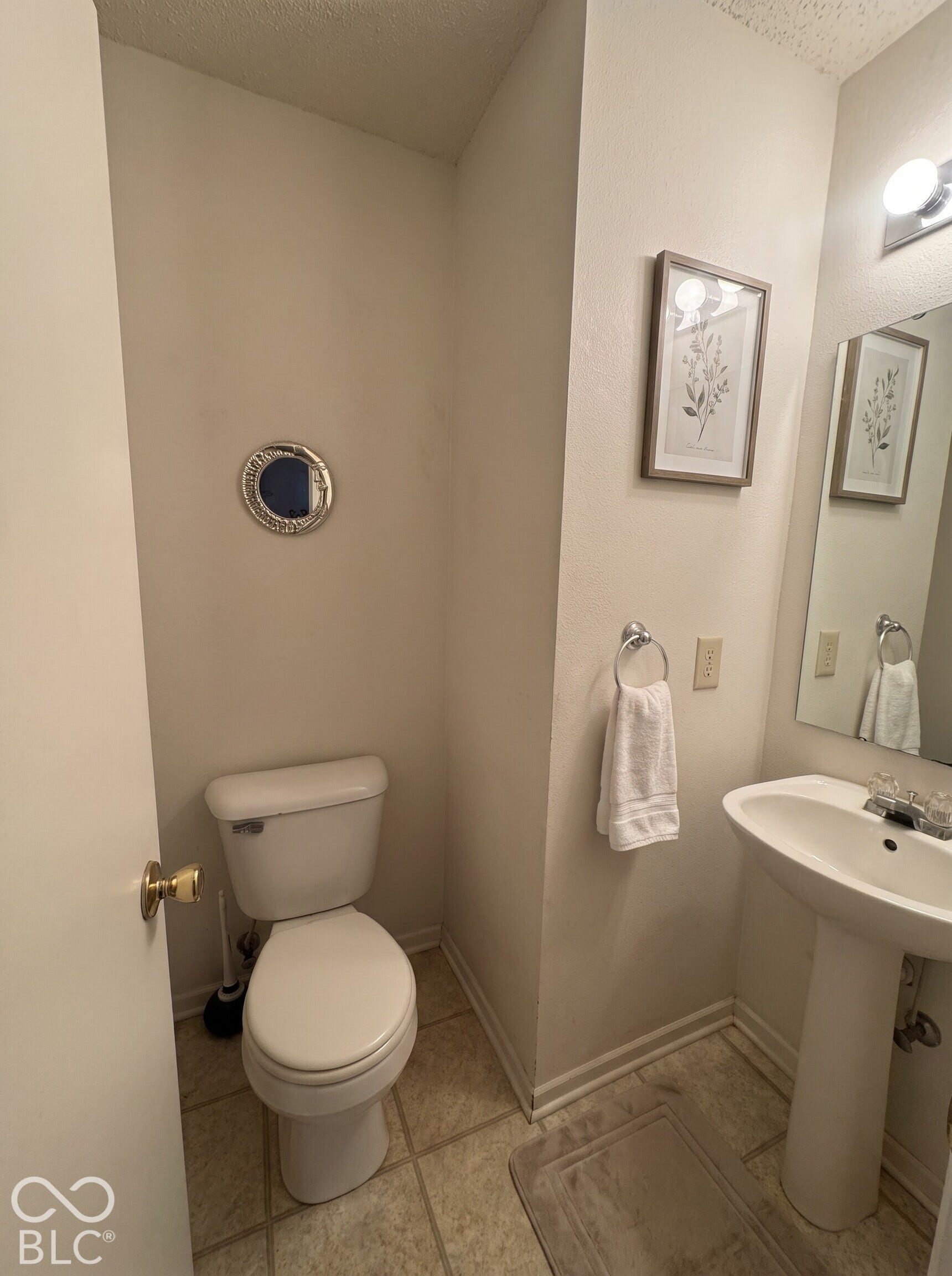 Virtually Altered - Powder room featuring a pedestal sink with chrome faucet, porcelain toilet, tiled flooring, and wall-mounted vanity lighting