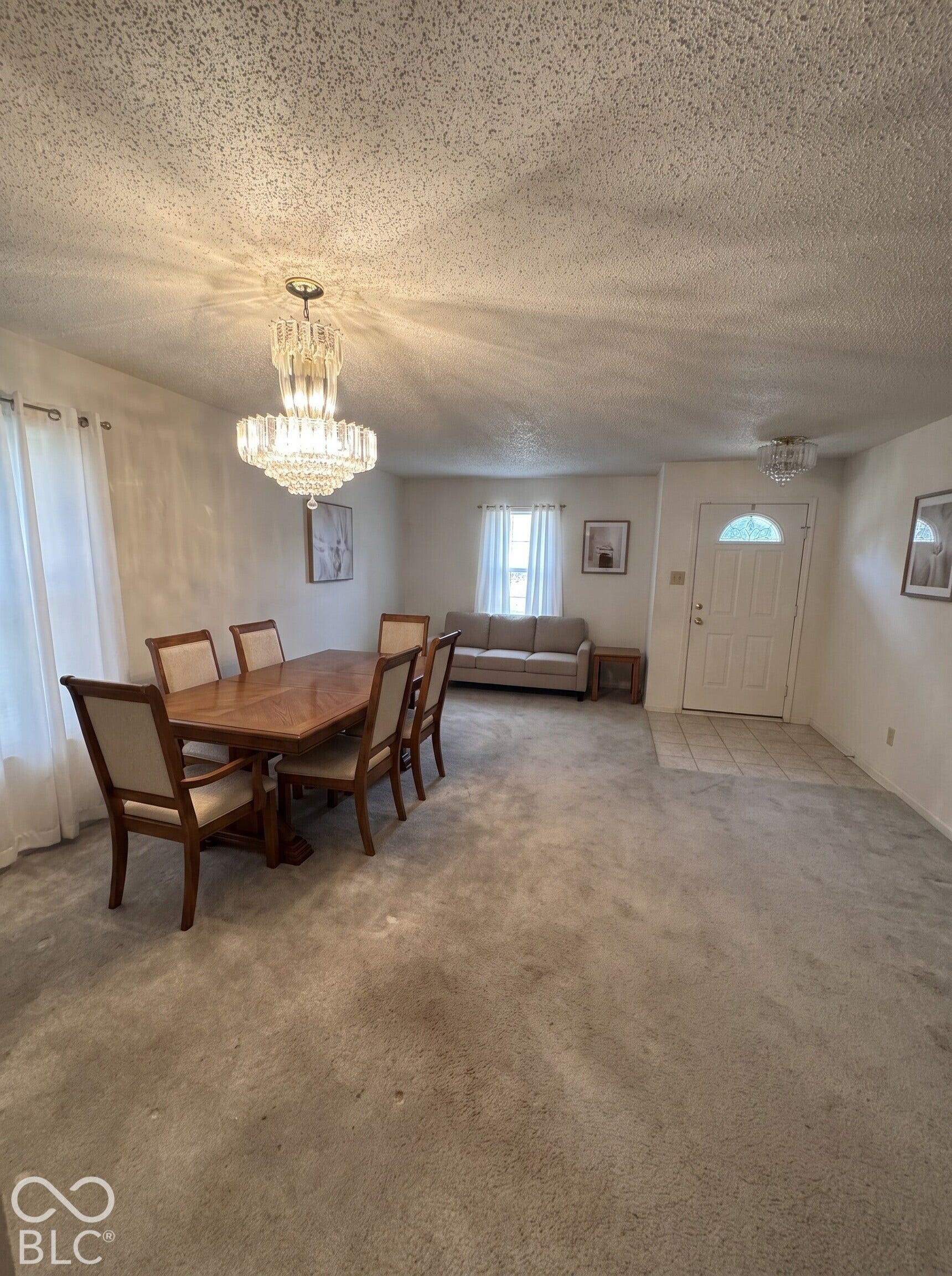 Virtually Altered - Open-concept area featuring a tiered crystal chandelier, secondary glass flush-mount lighting, and a neutral tile entryway