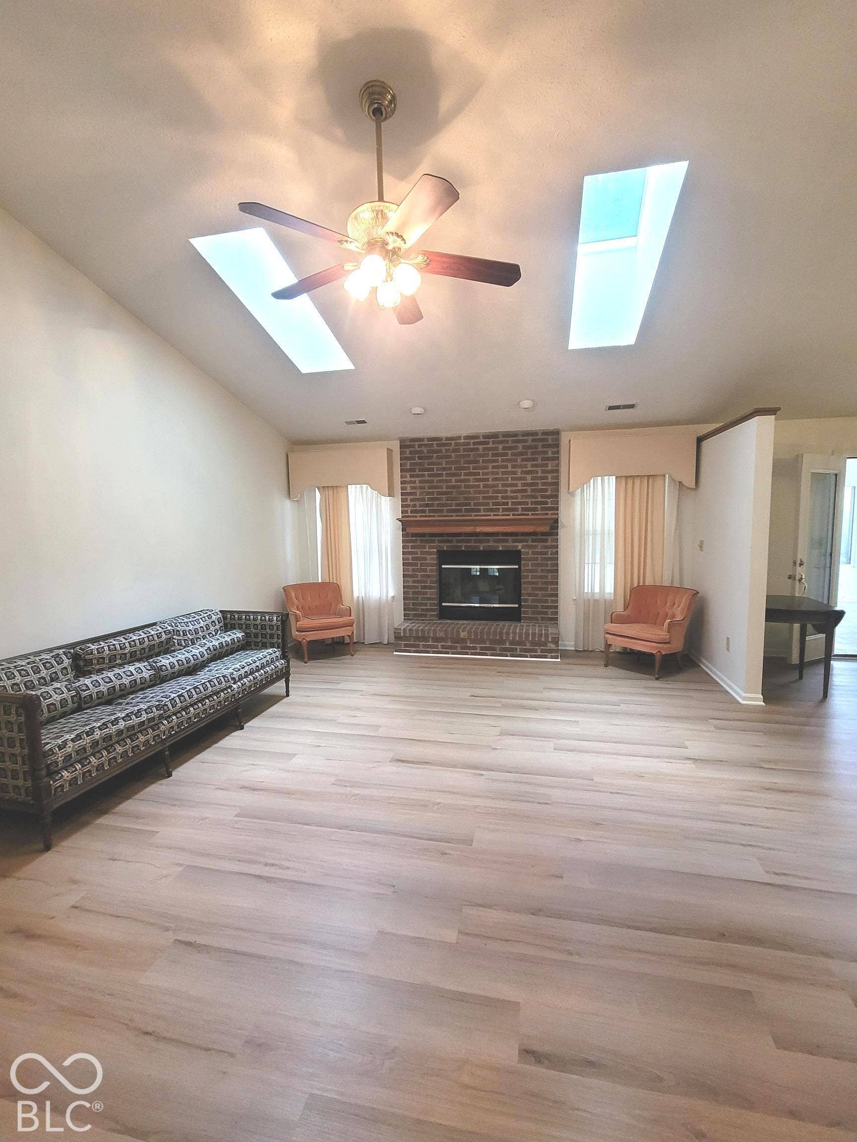 Vaulted ceilings with dual skylights, wood-finish flooring, brick fireplace with a wood mantel, and a central ceiling fan